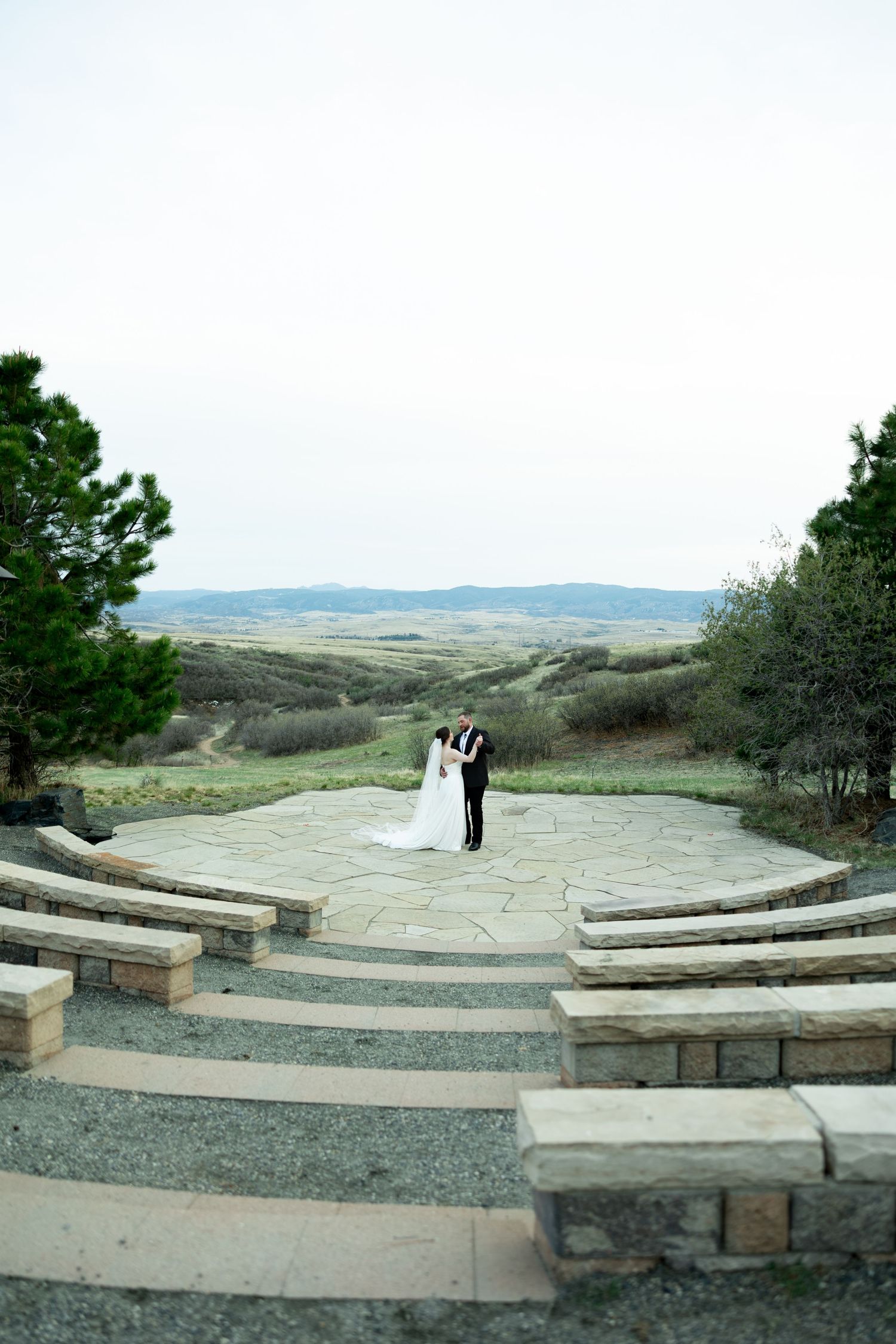 Bride and groom embrace on stone patio amphitheater overlooking scenic rolling hills landscape at outdoor wedding venue.