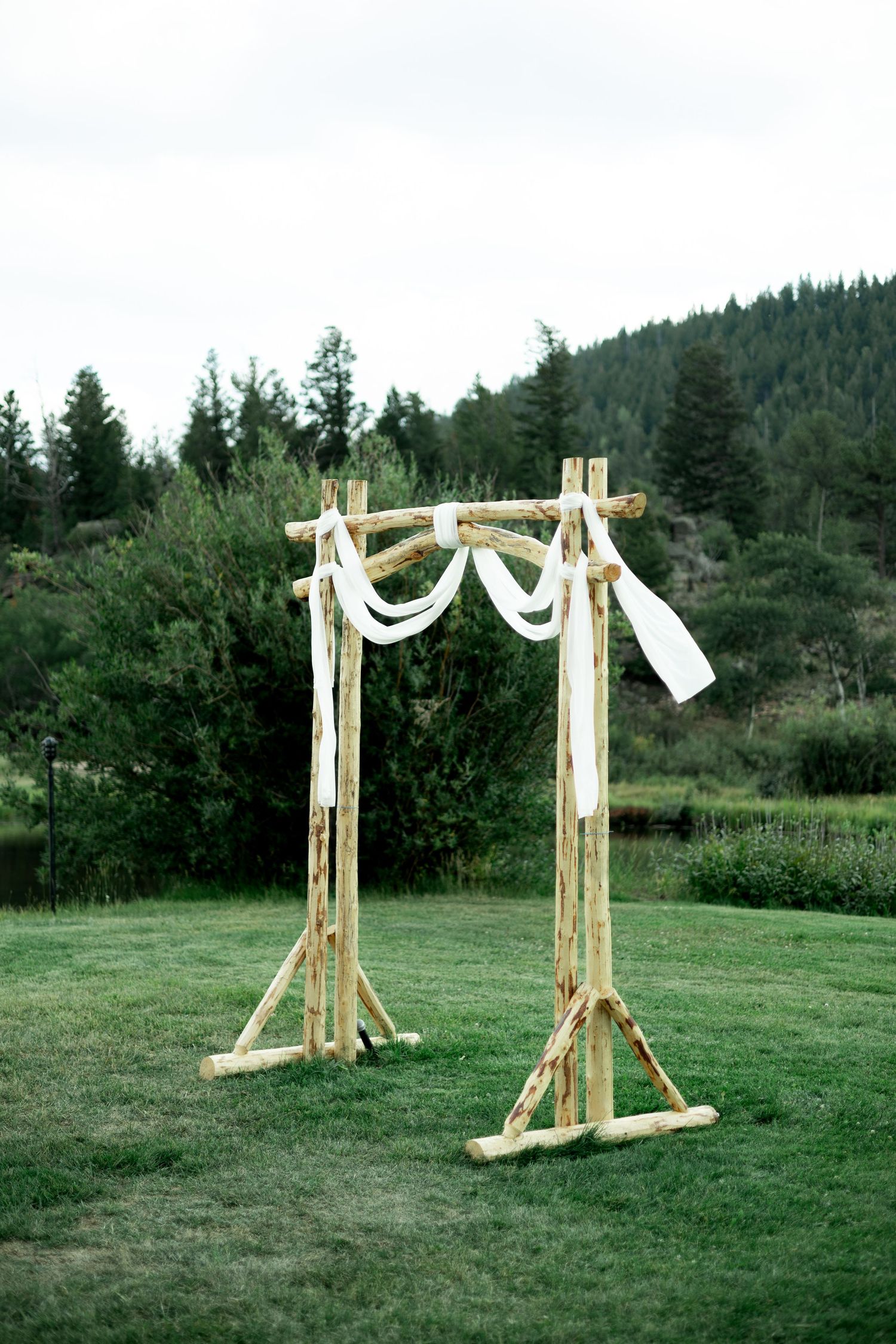 Wooden wedding arch structures with white fabric draping set up in a green field with forest backdrop.