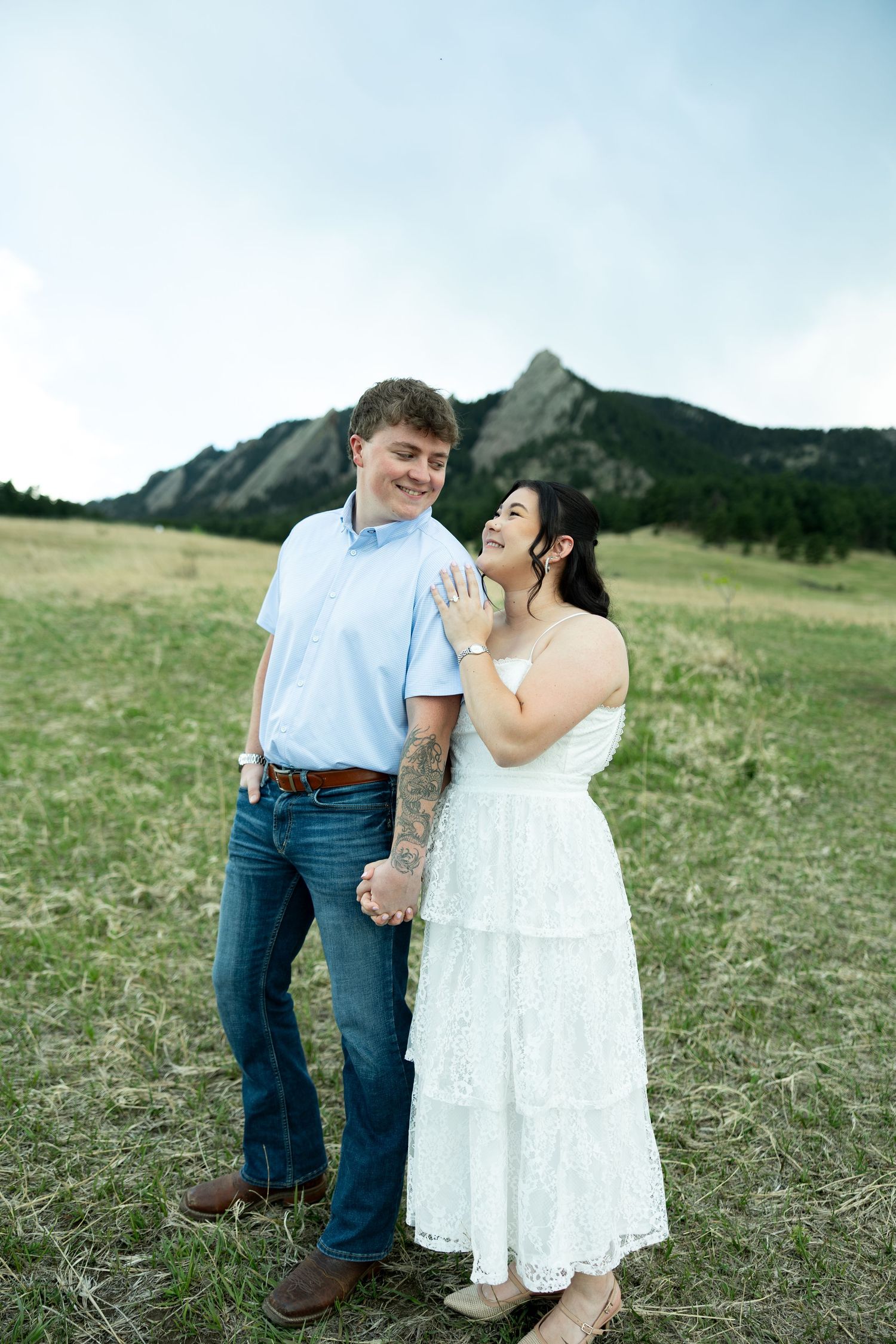Couple holding hands in a mountain meadow, woman in white lace dress showing engagement ring near Colorado Flatirons.