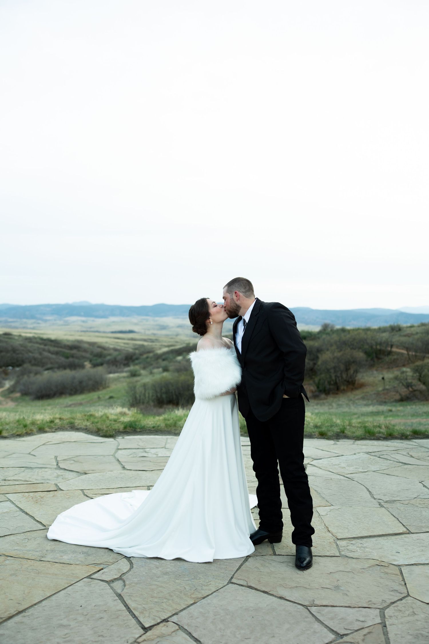 Bride and groom share a kiss on a stone terrace with scenic rolling hills in the background at their outdoor wedding.