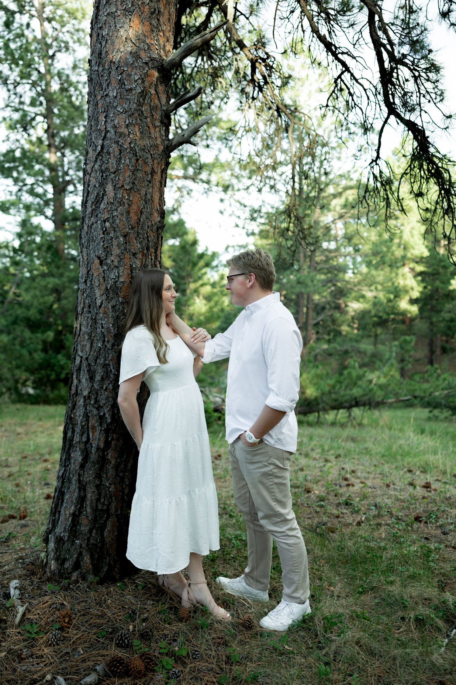 Couple in white outfits sharing a romantic moment by a pine tree in a lush green forest.