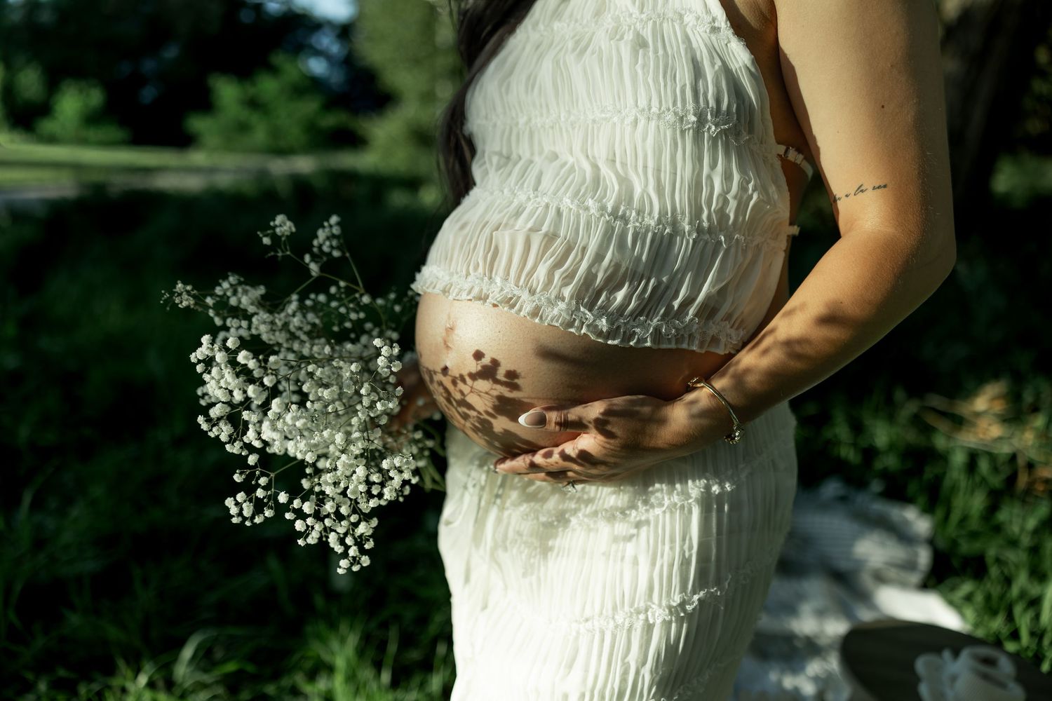 Pregnant woman in white fringe dress holding baby's breath flowers, cradling her bare bump outdoors.
