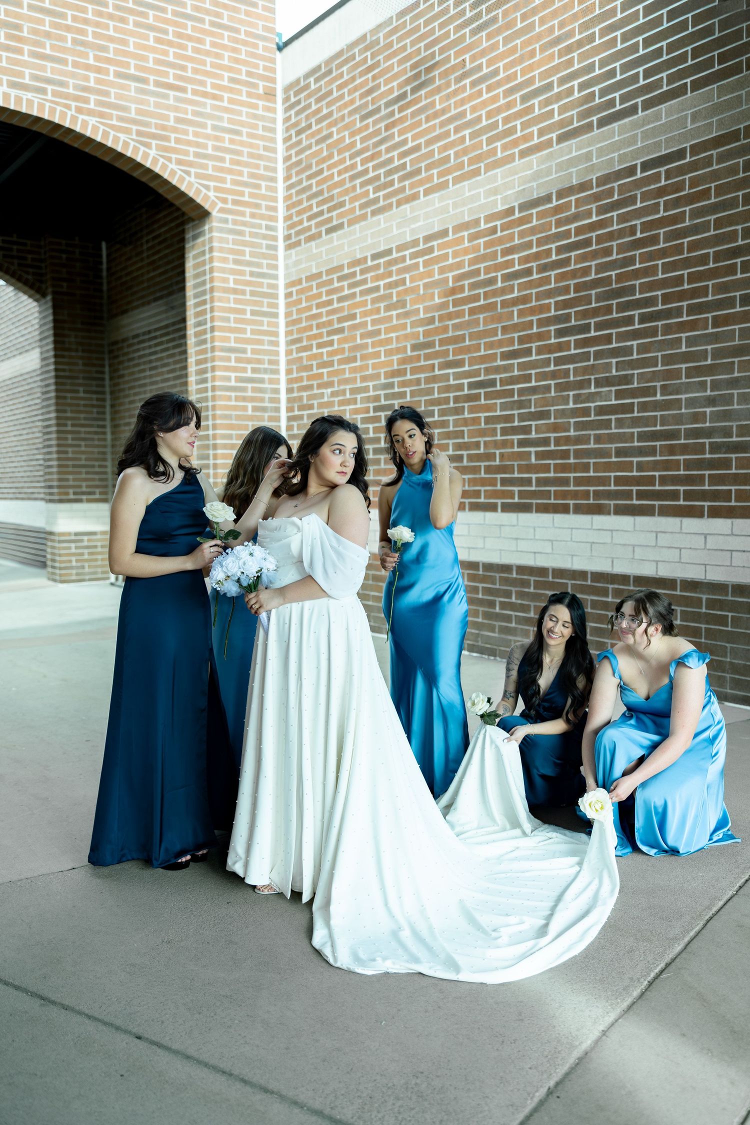 Bride in white gown with bridesmaids in blue satin dresses posing near brick wall outdoors.