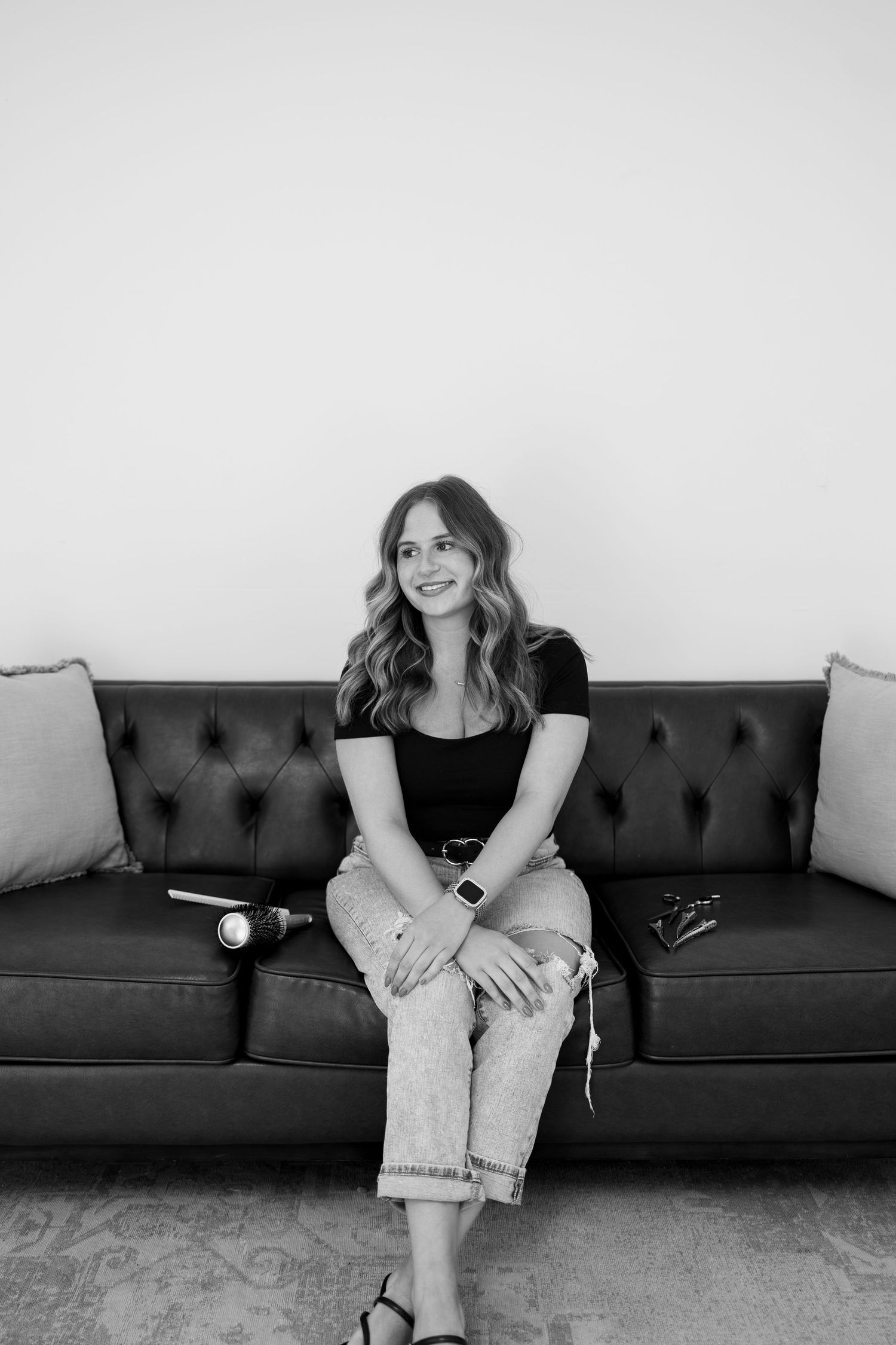Young woman with wavy hair sits smiling on a tufted leather sofa in a black-and-white portrait photo.