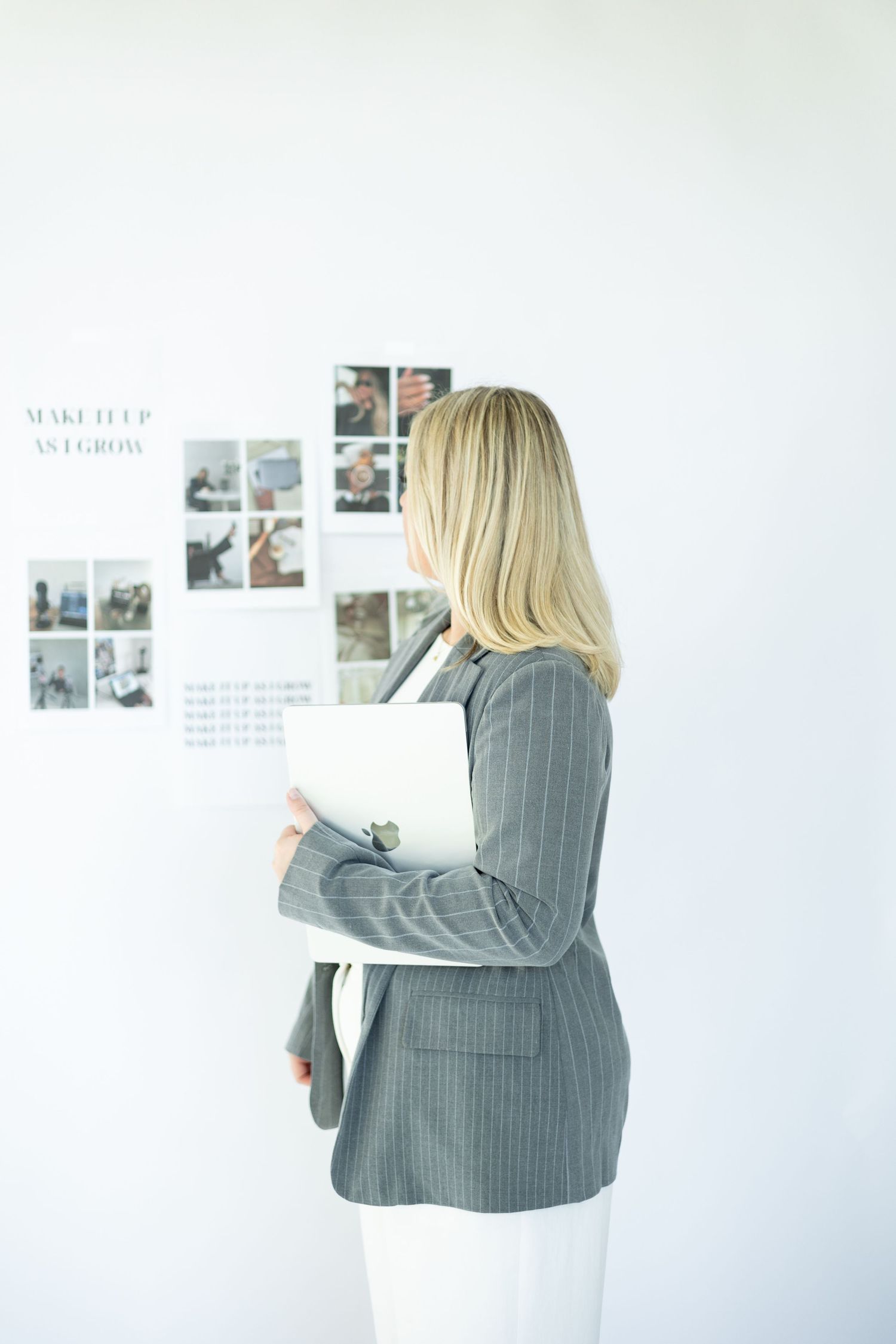 Professional in gray blazer reviews photos on wall while holding documents in minimalist office setting.