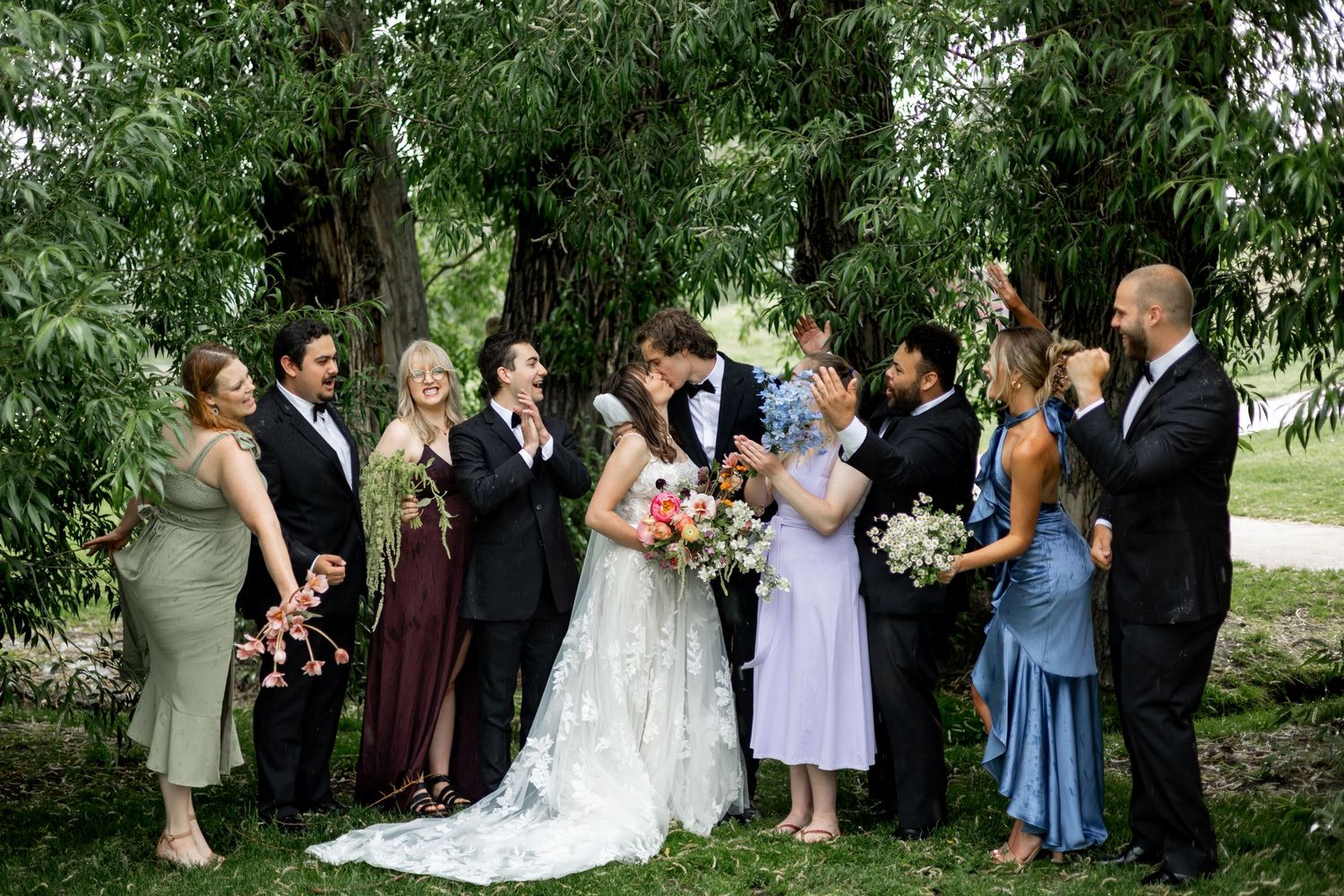 Wedding party in mixed-color formal attire poses together outdoors under trees.