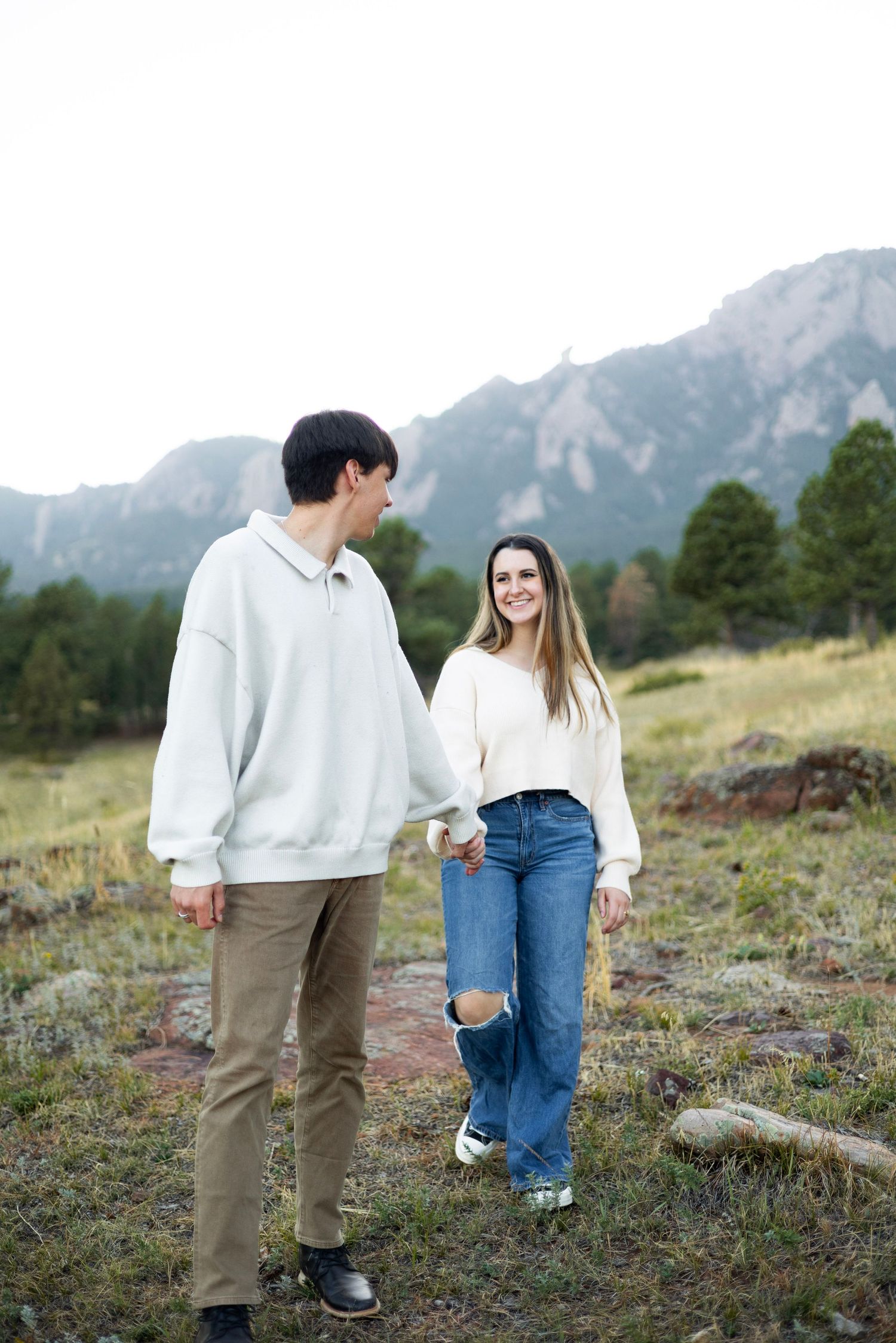 A couple in matching white sweaters stands together on a mountain hillside.