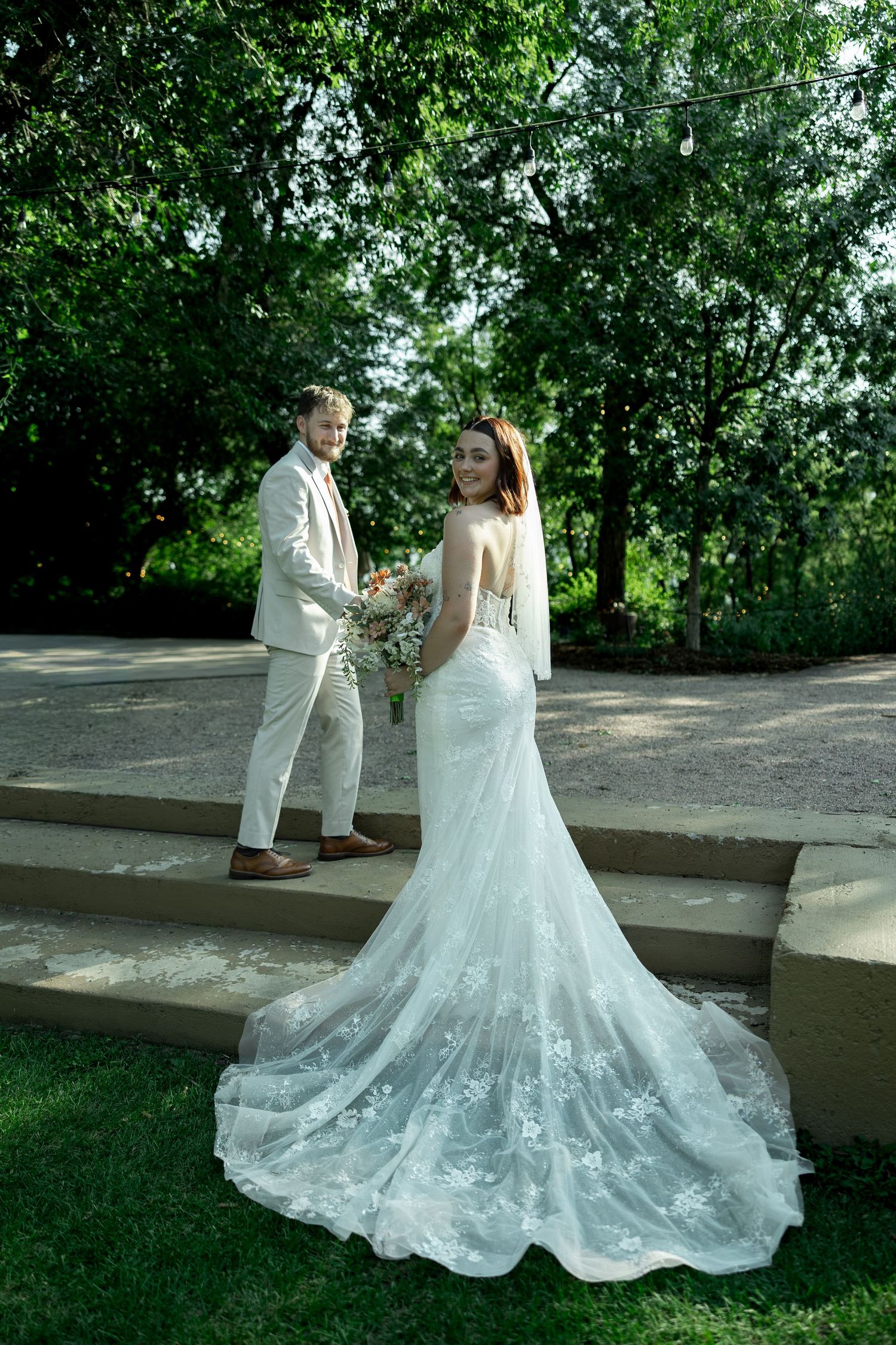 Bride in lace mermaid gown with train smiles over shoulder as groom in beige suit stands on stone steps in garden.