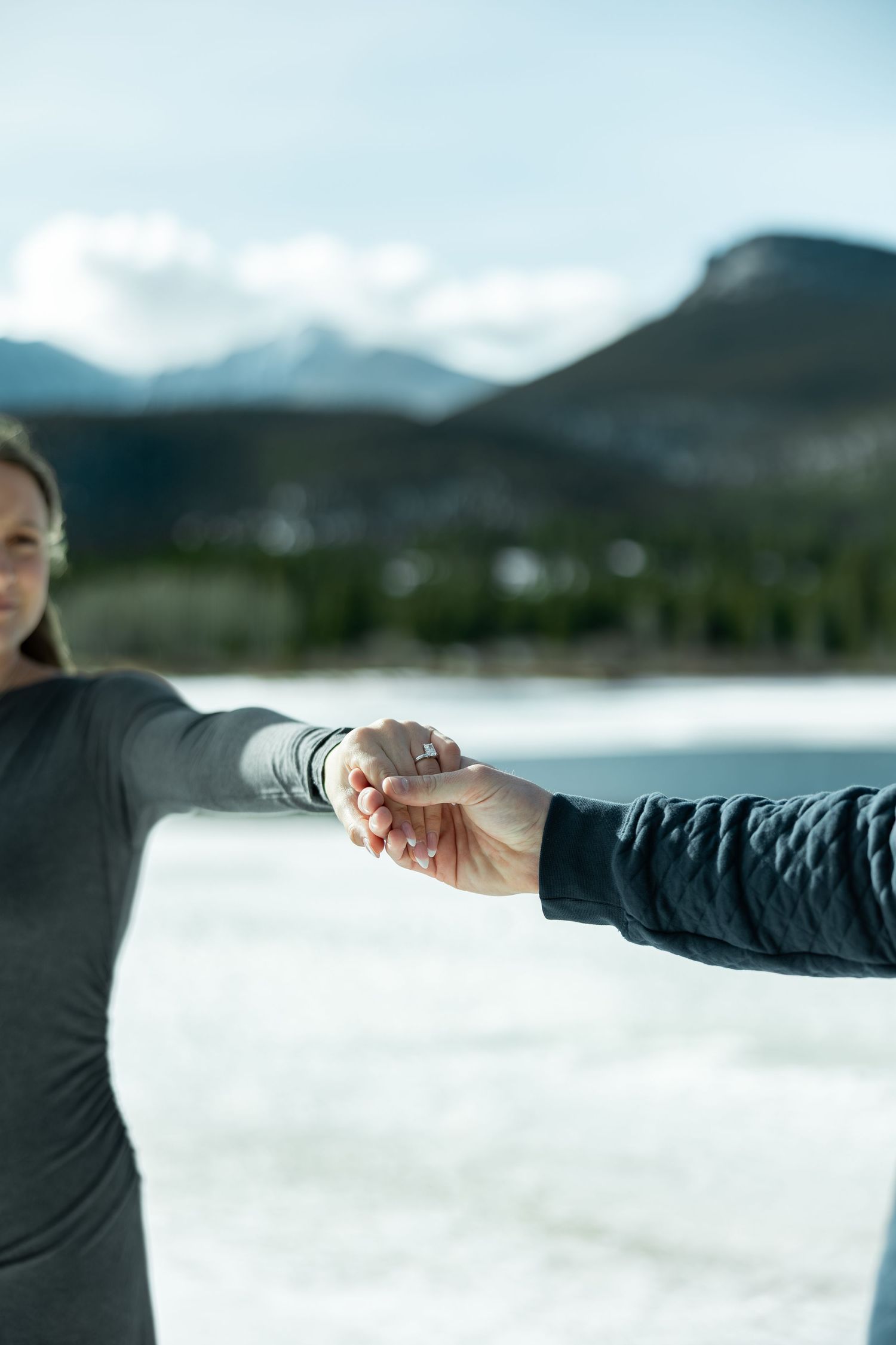 Two people holding hands against a scenic mountain landscape with snow and cloudy sky in the background.