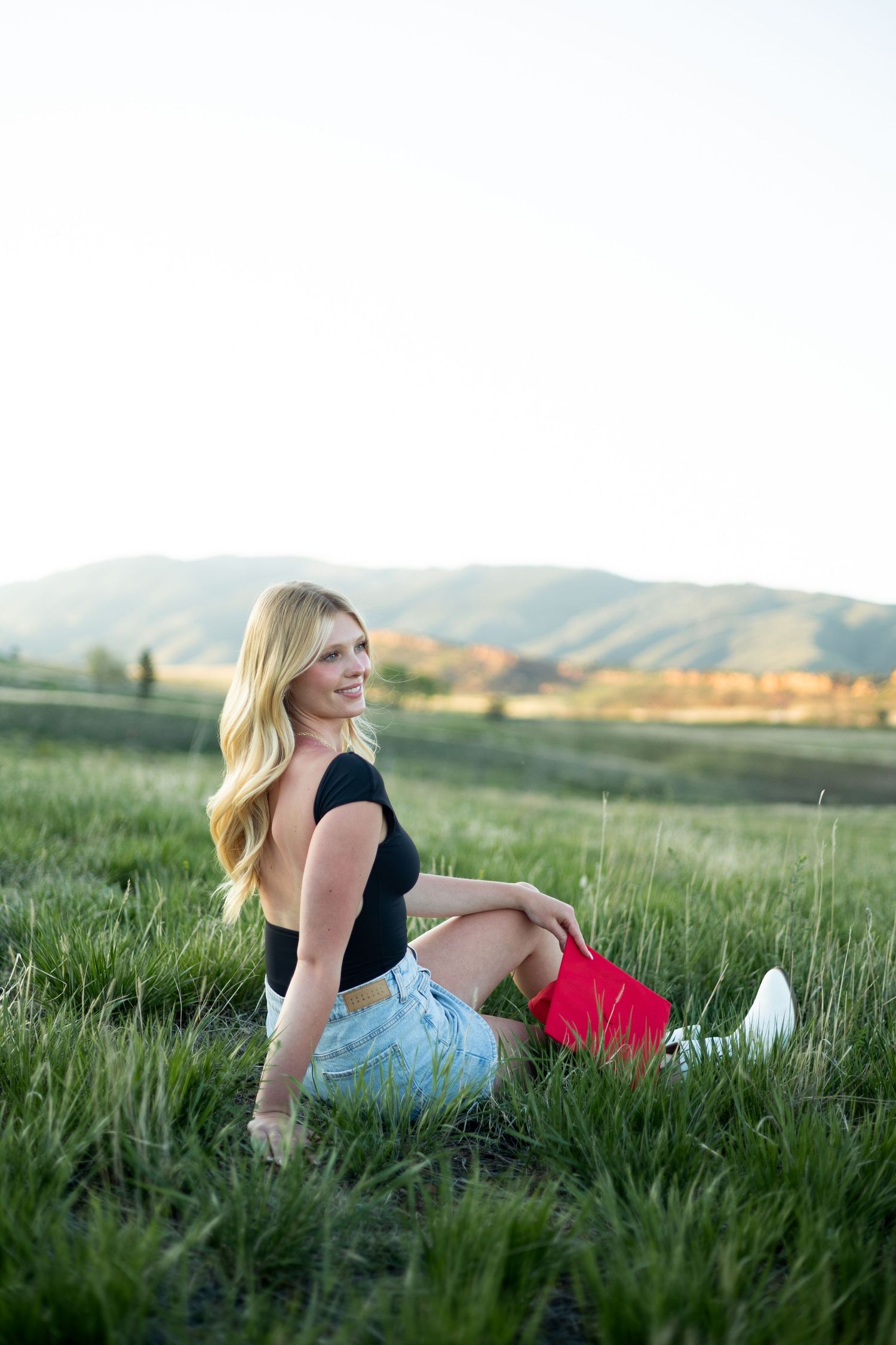 Blonde woman sitting in green grass field wearing black crop top and denim shorts with mountains in background.