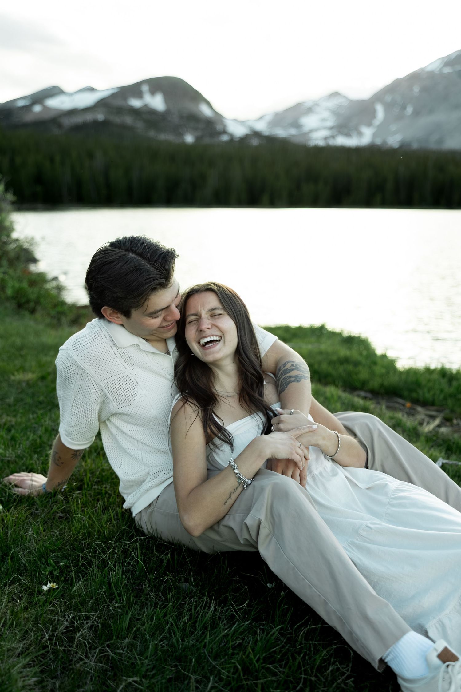 A laughing couple sits on grass by a mountain lake, the man holding the woman as she throws her head back in joy.
