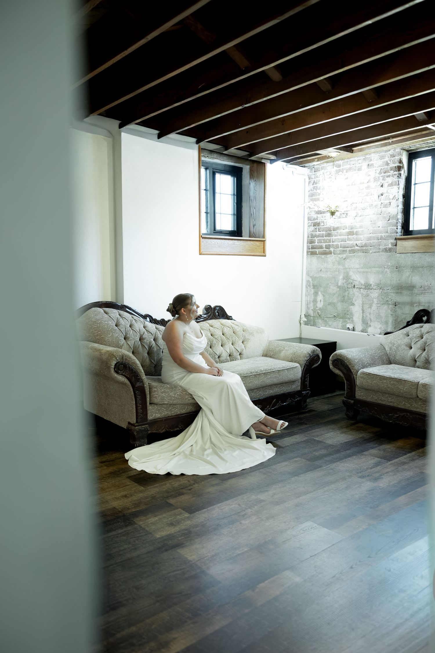 Bride in white gown sitting elegantly on a tufted sofa in a rustic room with hardwood floors and exposed ceiling beams.
