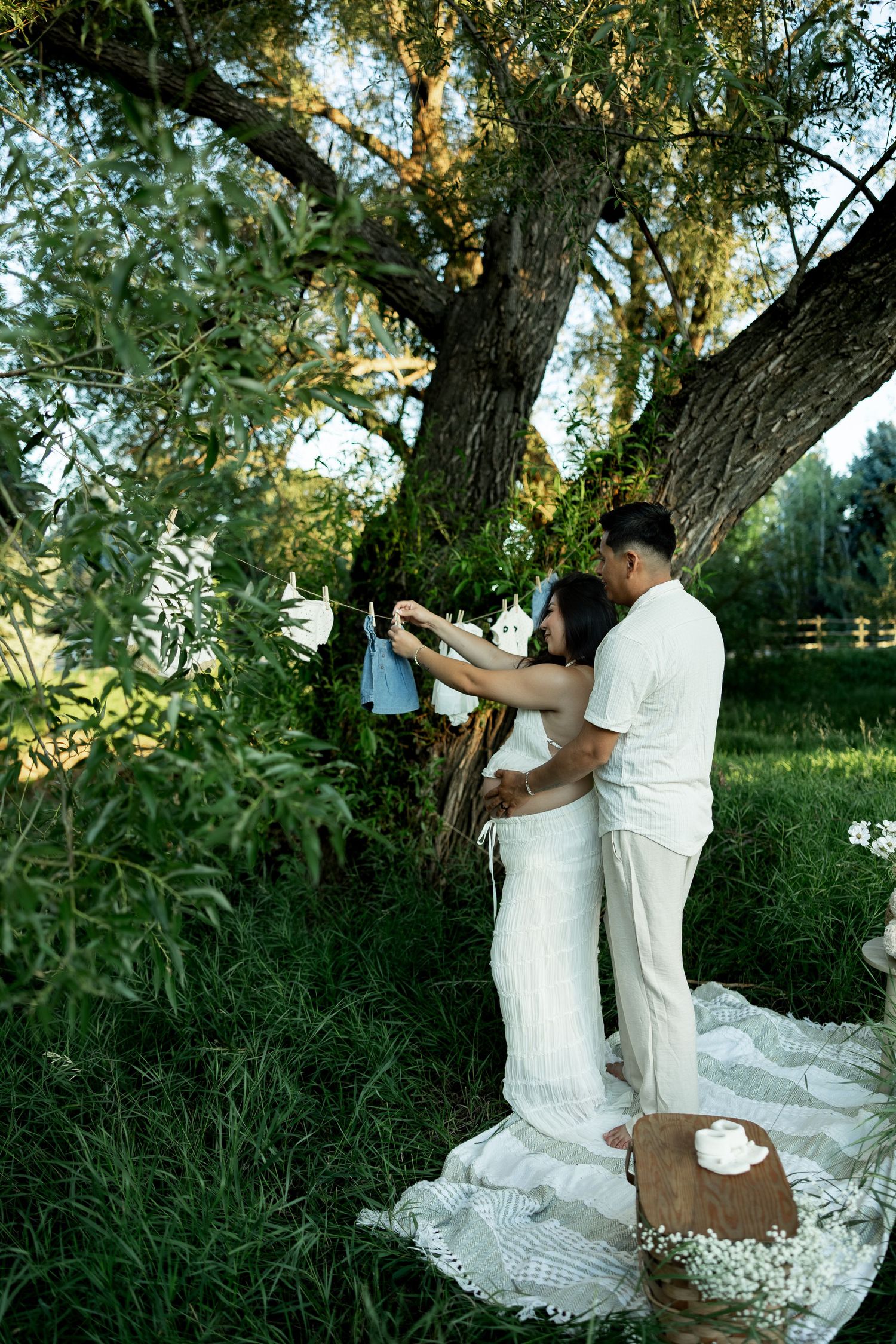 Pregnant couple in white outfits hanging baby clothes on a tree branch during an outdoor maternity photoshoot.
