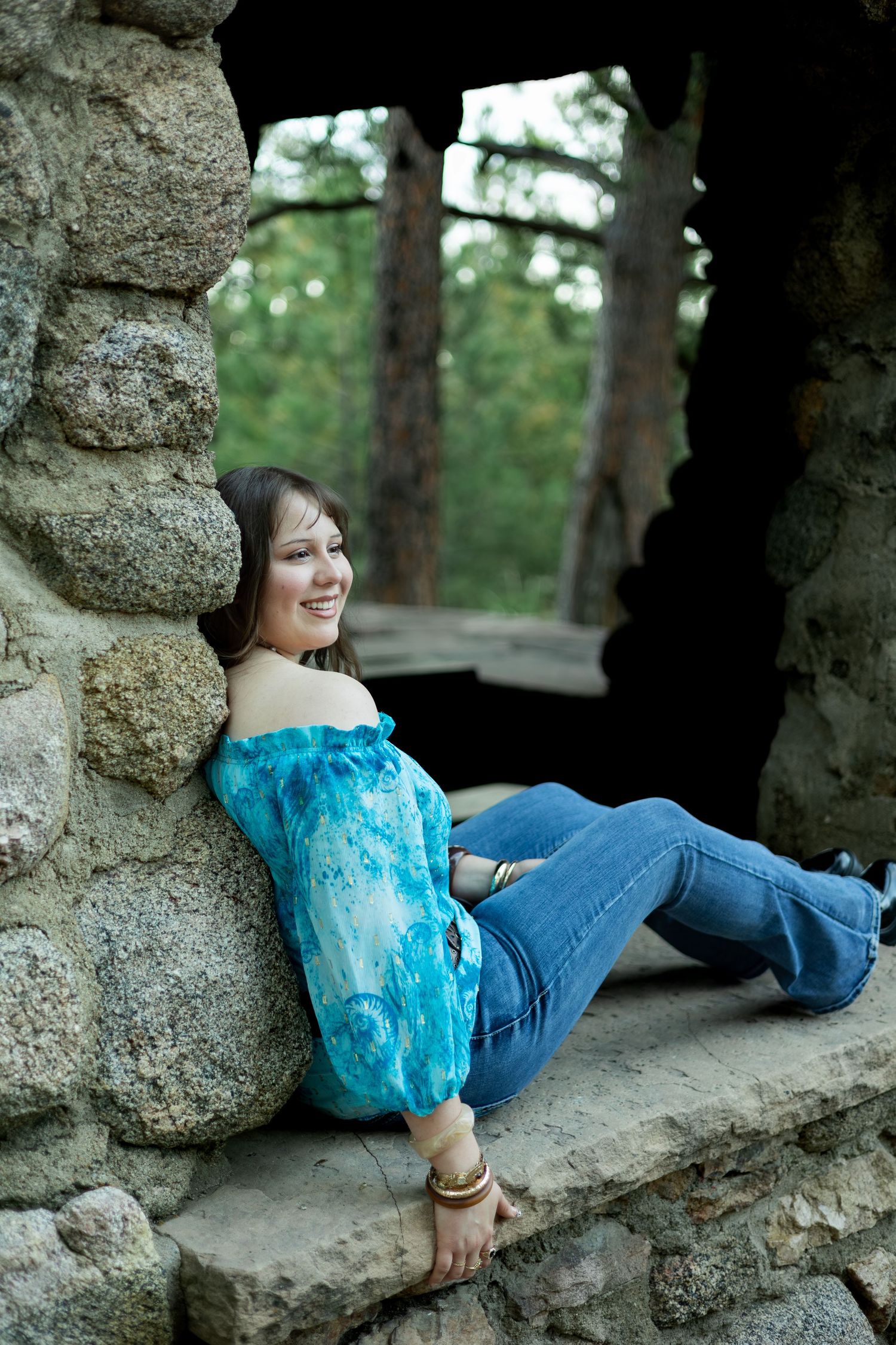 Person in turquoise top and jeans relaxes against stone wall in outdoor nature setting.