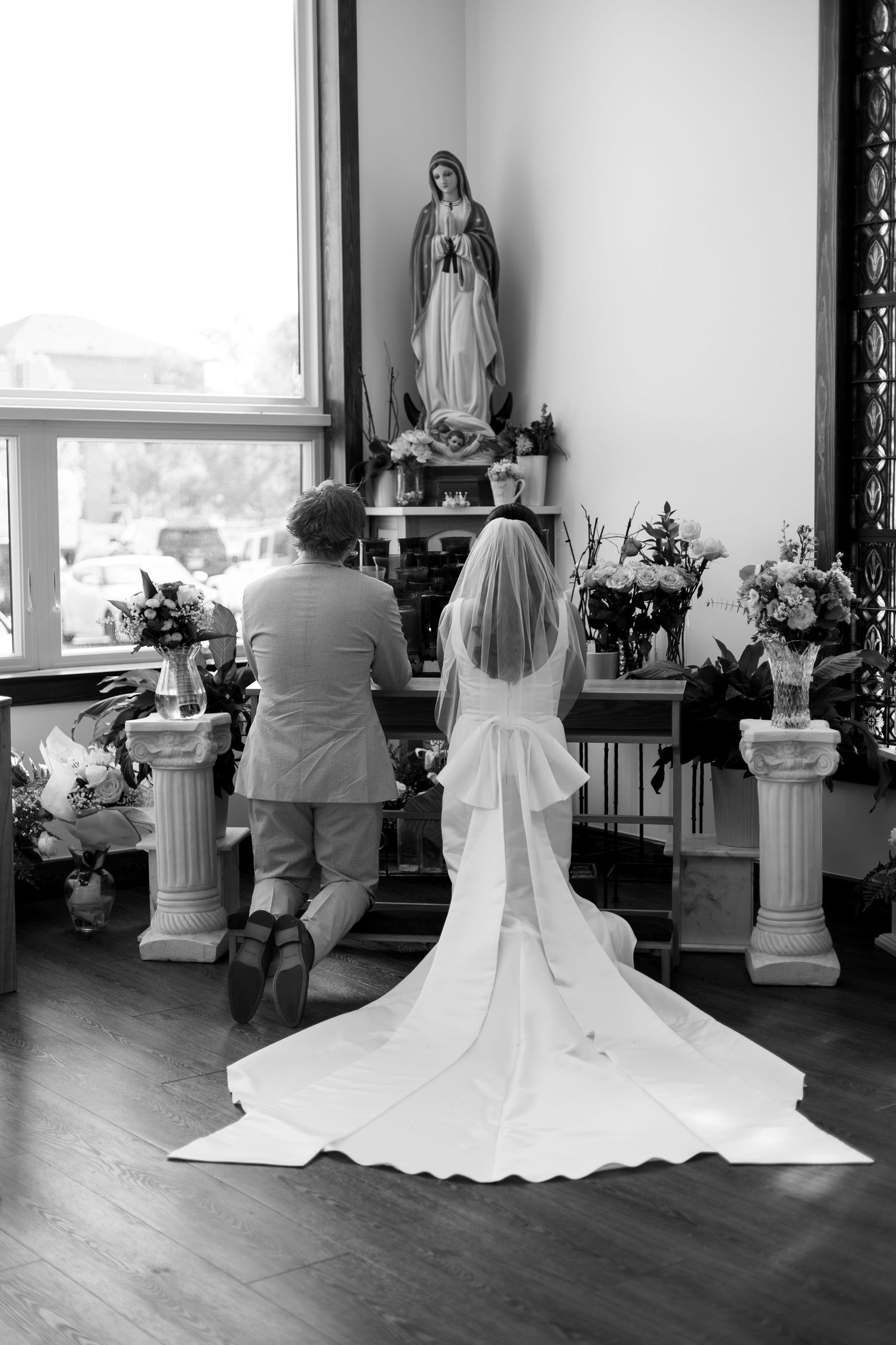 Black and white photo showing a bride in a flowing white dress with long veil kneeling before a religious statue and flowers.