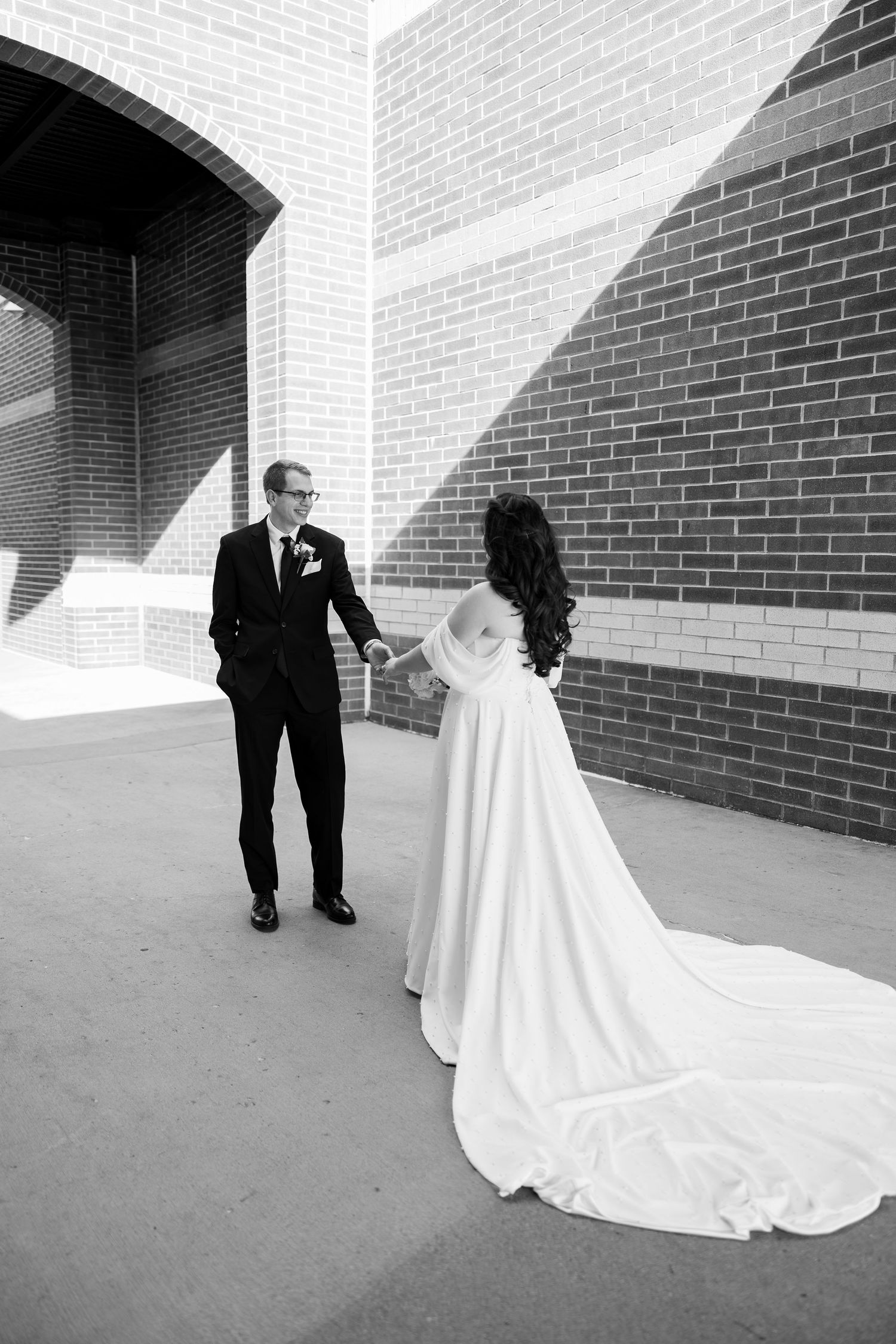 Black and white photo of bride and groom holding hands near brick wall.