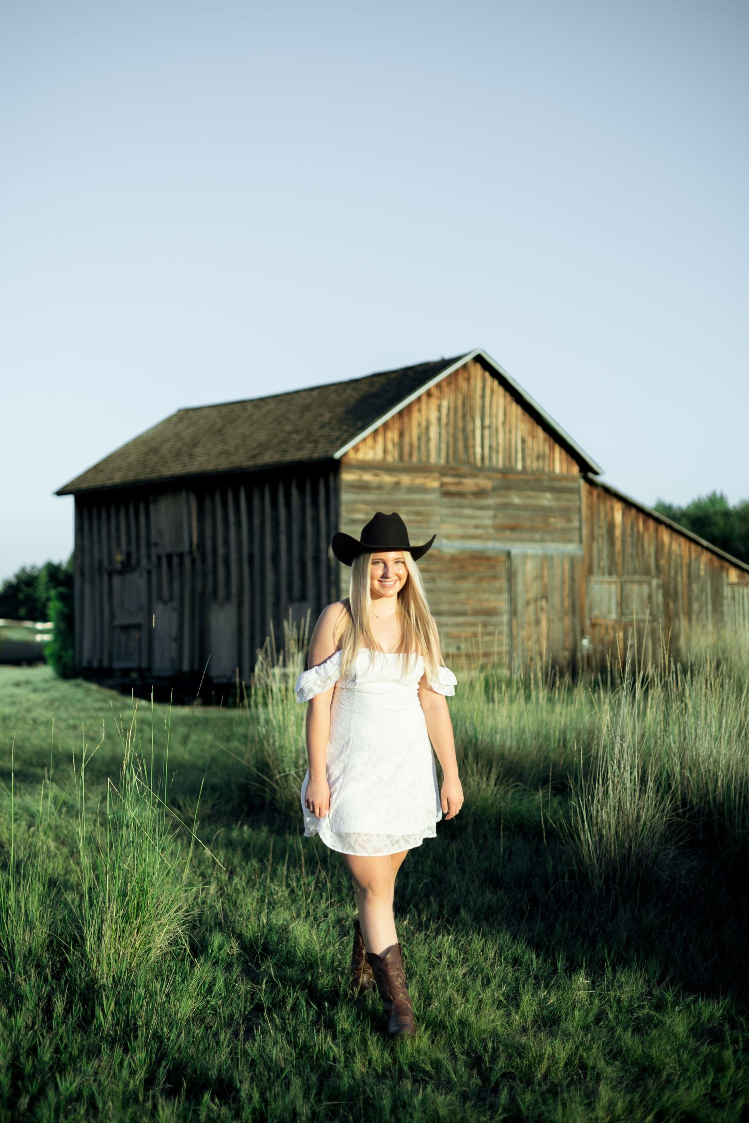 Smiling blonde woman in white dress, black cowboy hat, and brown boots standing in green field near rustic wooden barn.