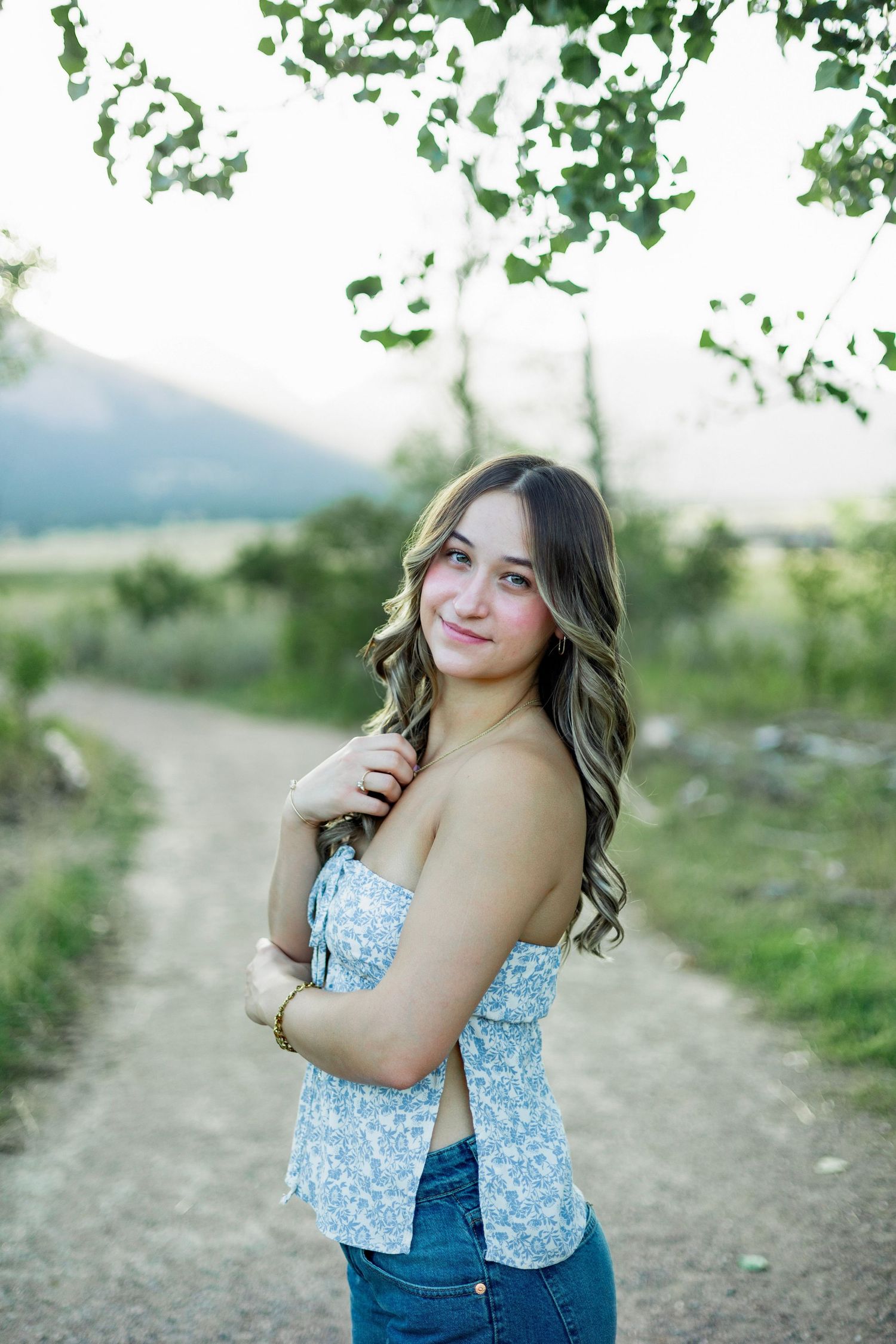 Young woman in floral tube top and jeans smiling on a rural dirt path surrounded by green trees and mountains.