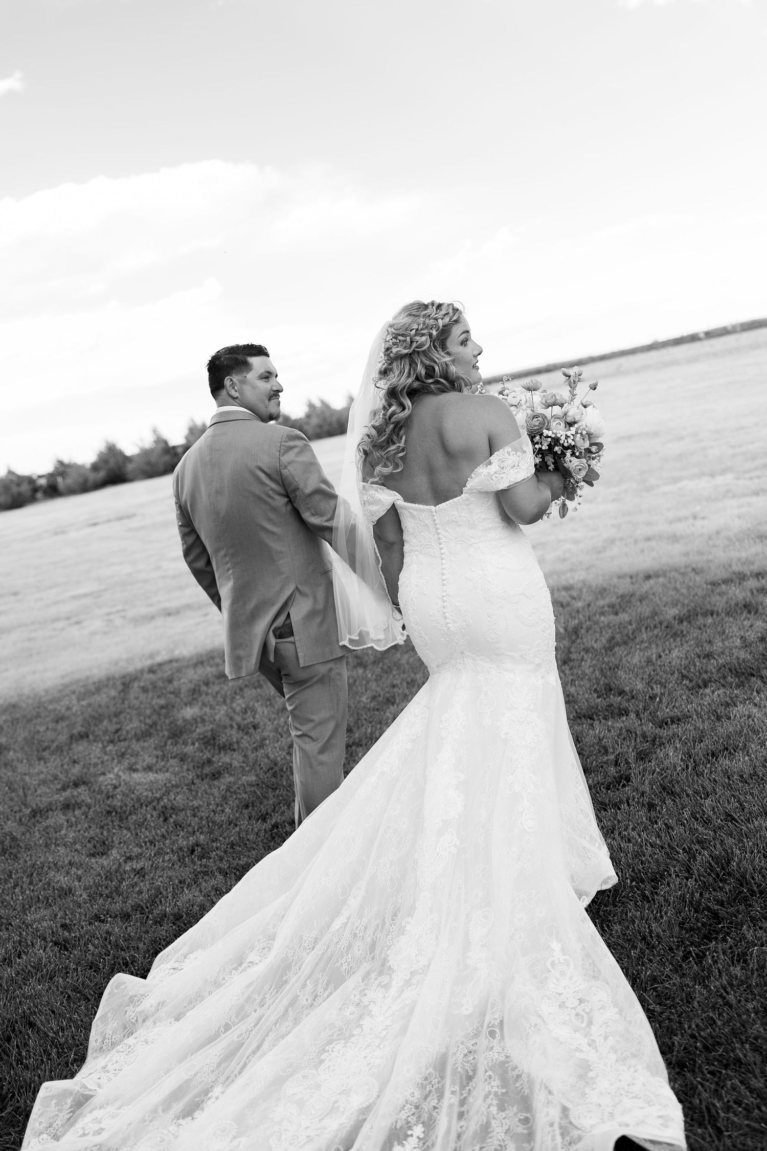 Black and white photo of bride in lace mermaid gown holding bouquet walking with groom in field.