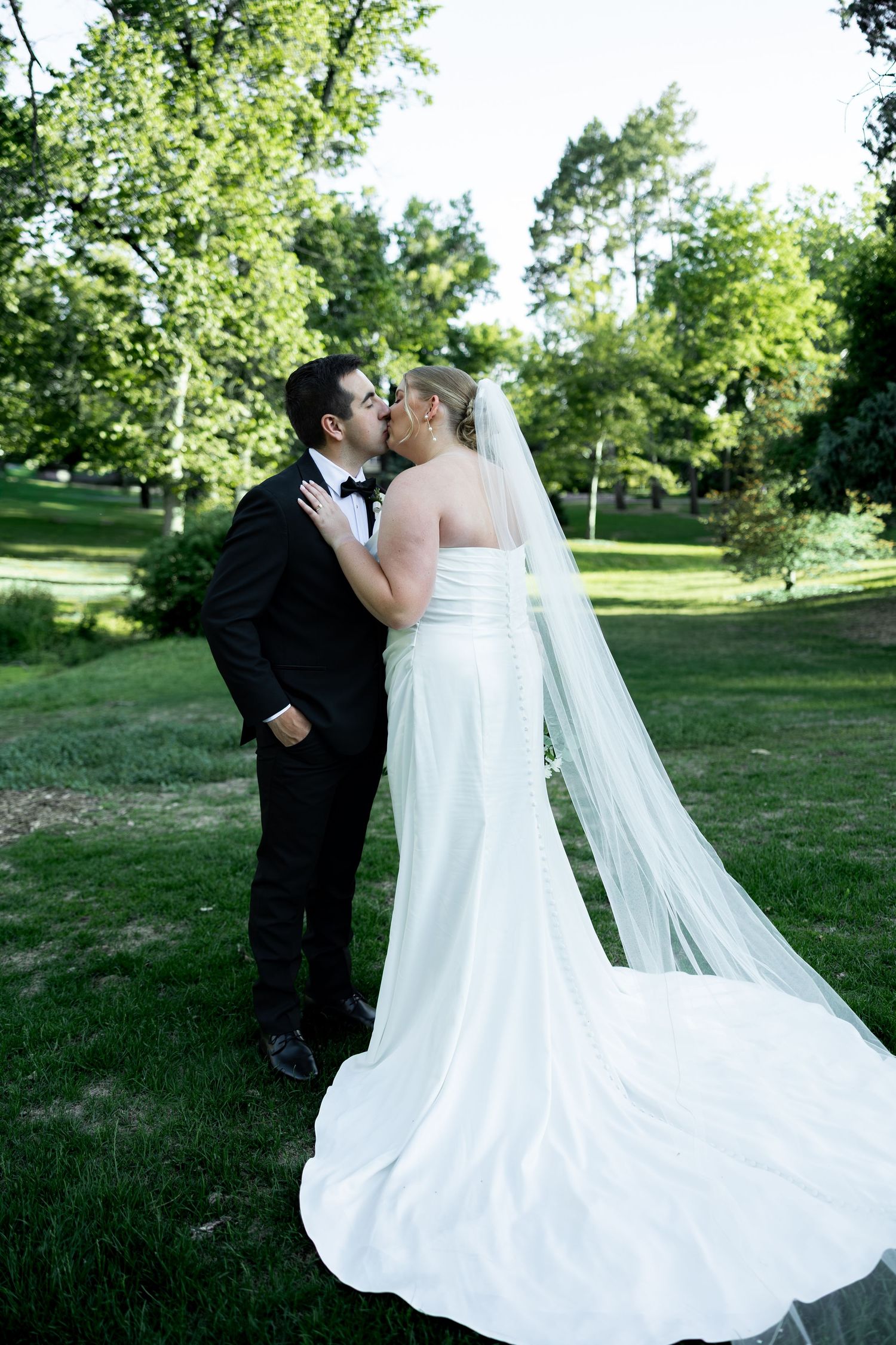 Bride and groom share a romantic kiss outdoors in a lush green park, bride wearing a white gown with long veil.