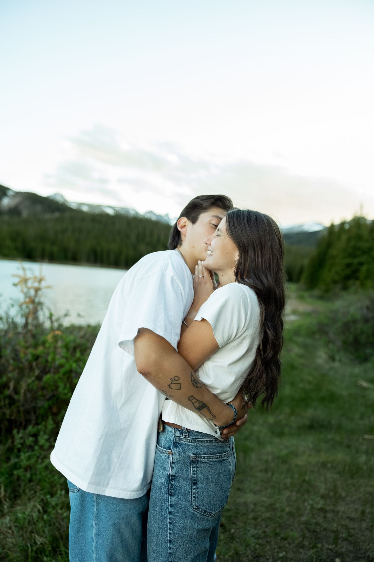 Two women in white shirts and jeans share a romantic embrace outdoors near a lake with mountain views.