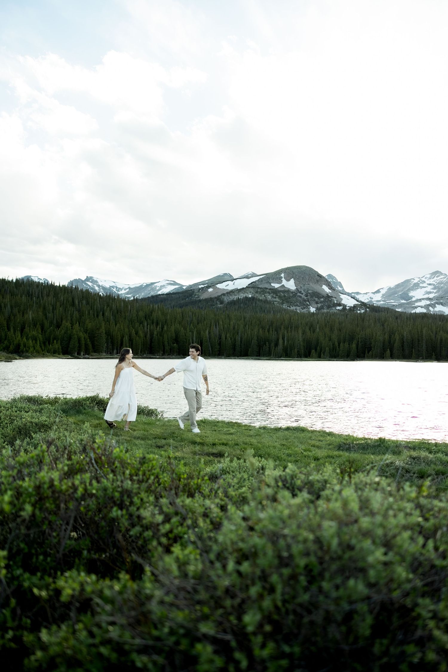 Couple in white holding hands by a mountain lake with snow-capped peaks and evergreen forest backdrop.