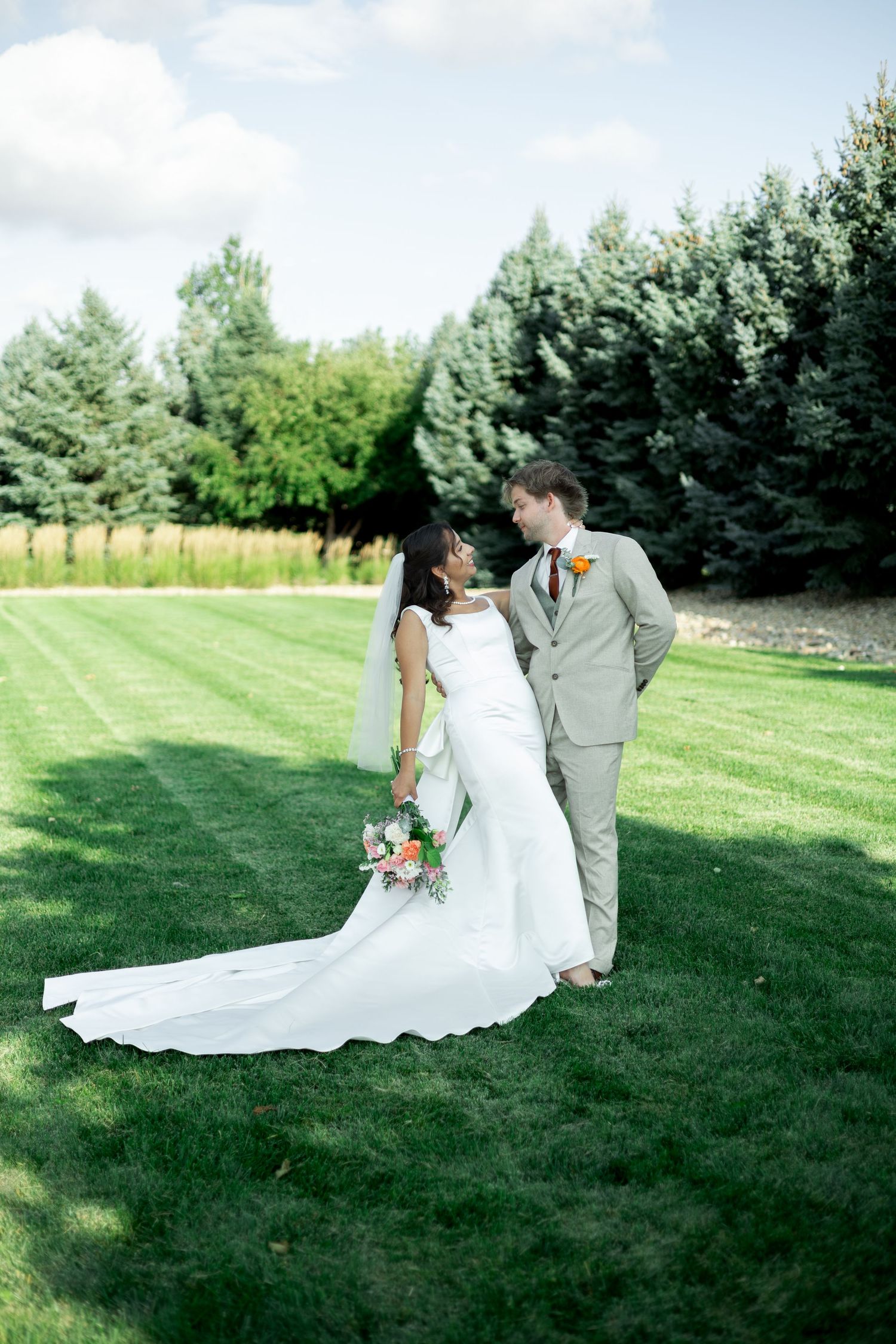 Bride and groom share a romantic moment on lush green lawn, she holds a colorful bouquet, evergreen trees in background.