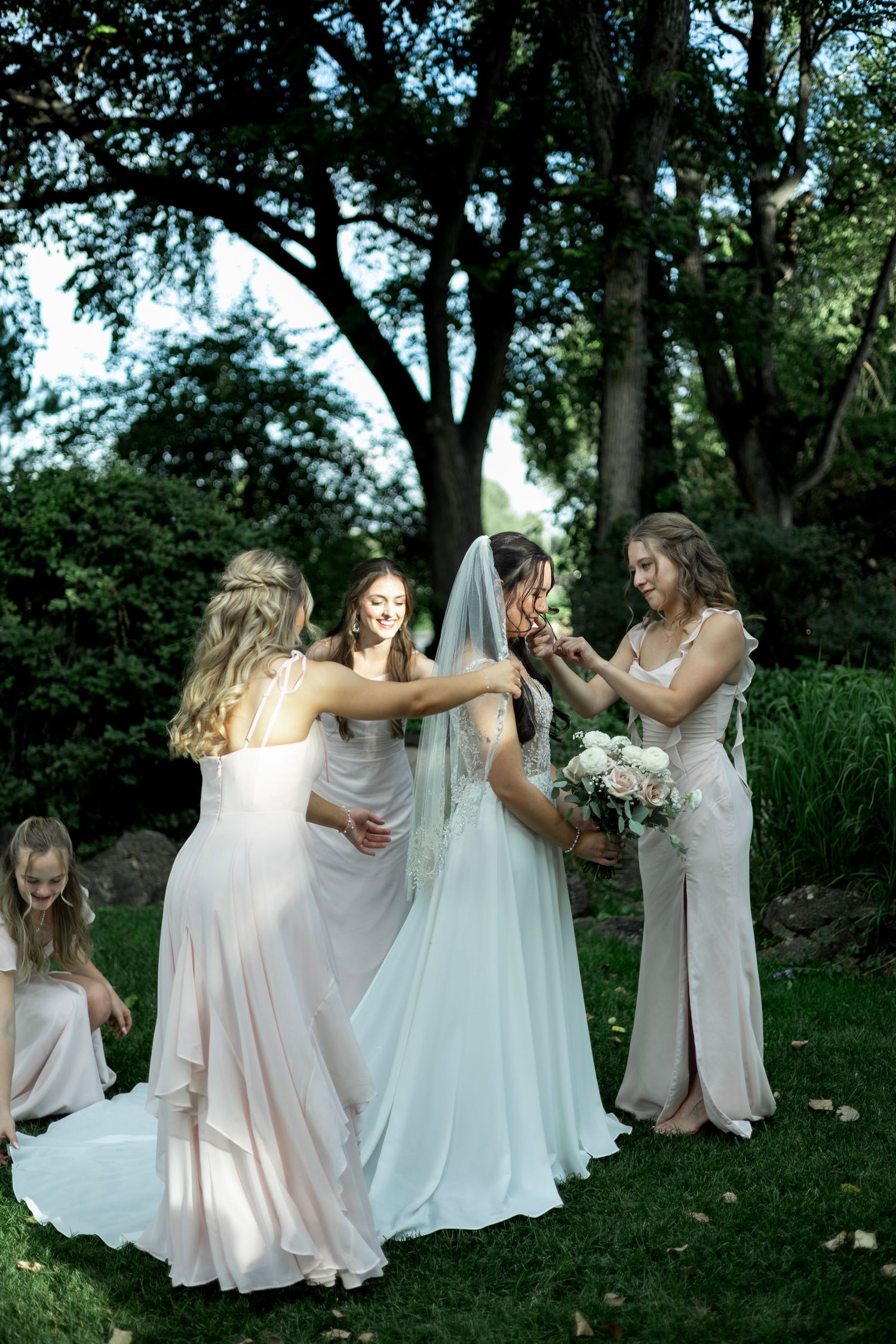 Bridesmaids in pale gowns celebrate with the bride on a green lawn surrounded by trees.