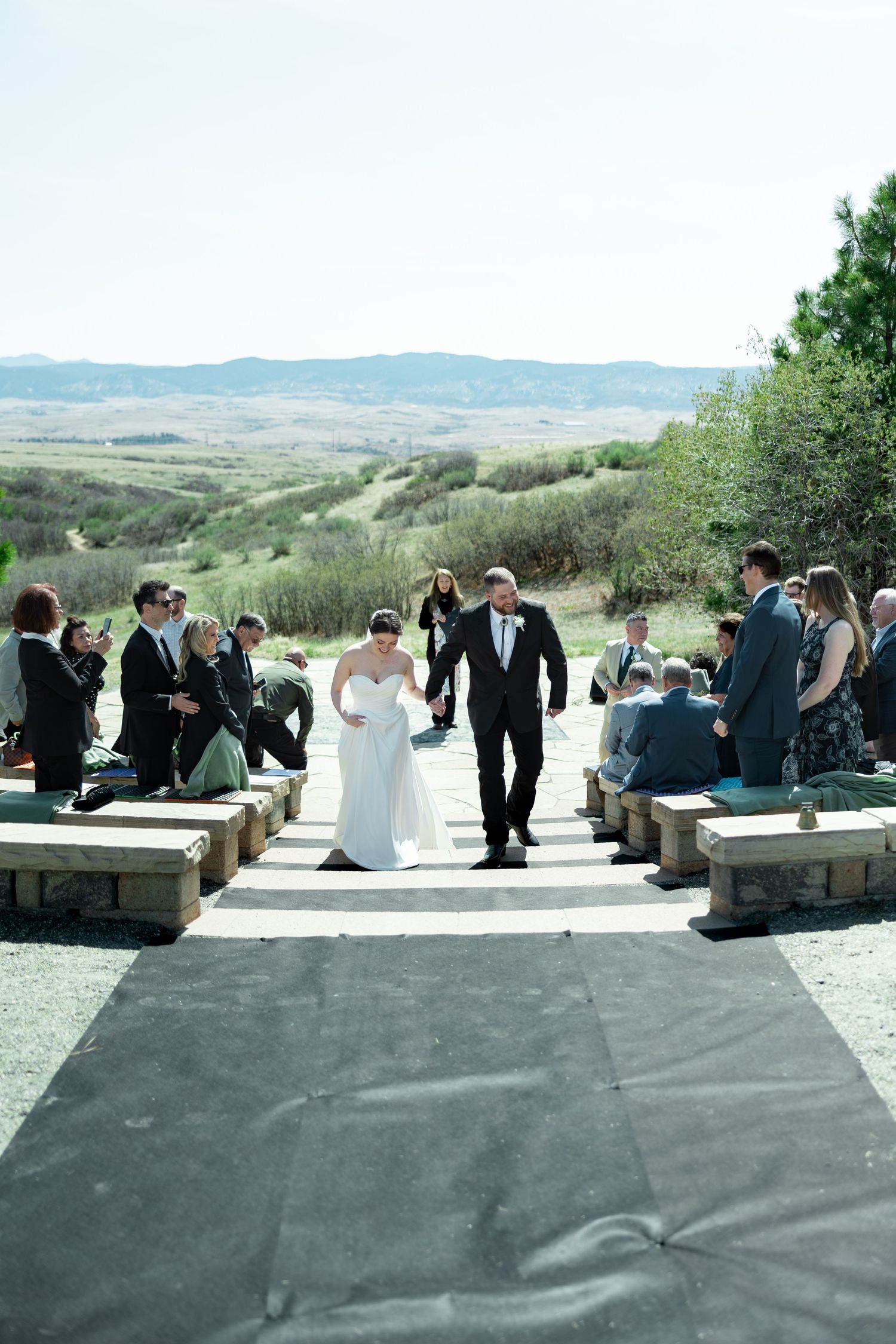 Bride and groom walk down outdoor stone aisle after ceremony with scenic rolling hills in background.