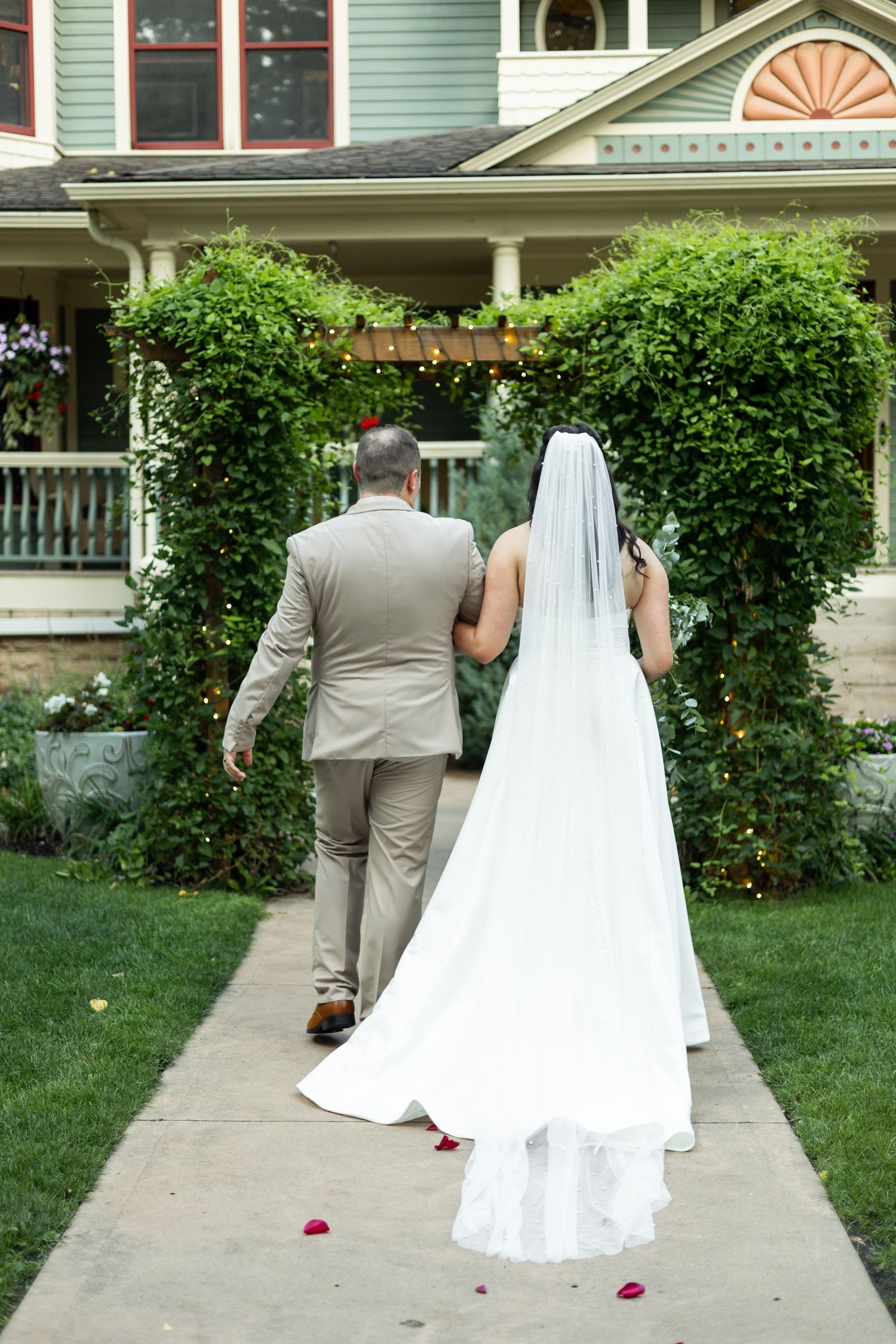 A couple in wedding attire walks down a garden path towards a Victorian-style house.