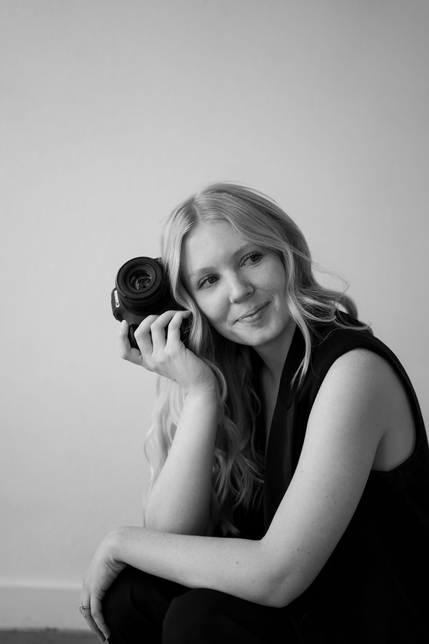 Black and white portrait of a photographer holding a camera while posing in a sleeveless black top.