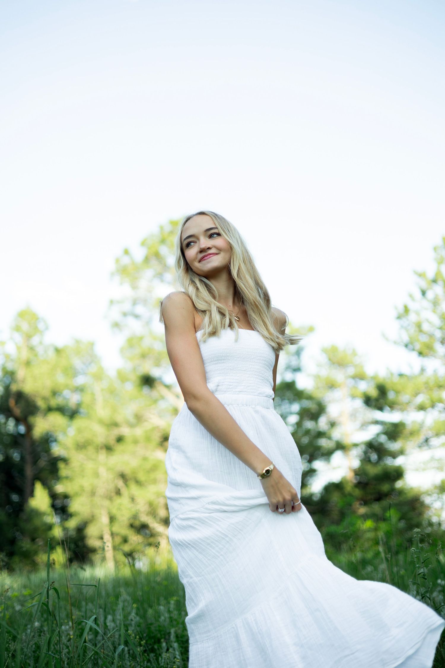 A person in a flowing white dress stands in a sunlit meadow surrounded by green trees.