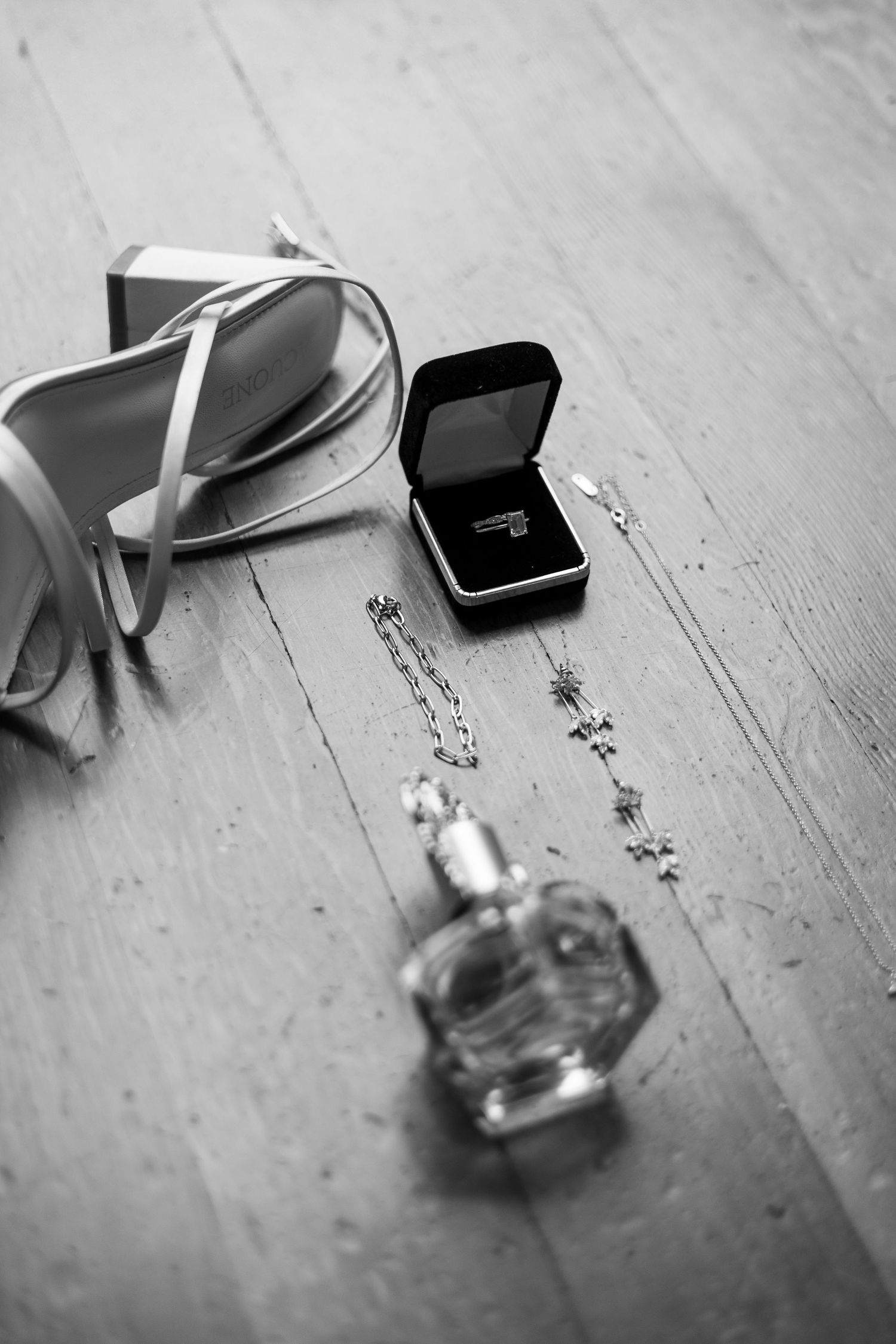 Wedding details including strappy sandals, ring box, and perfume bottle on wooden floor.