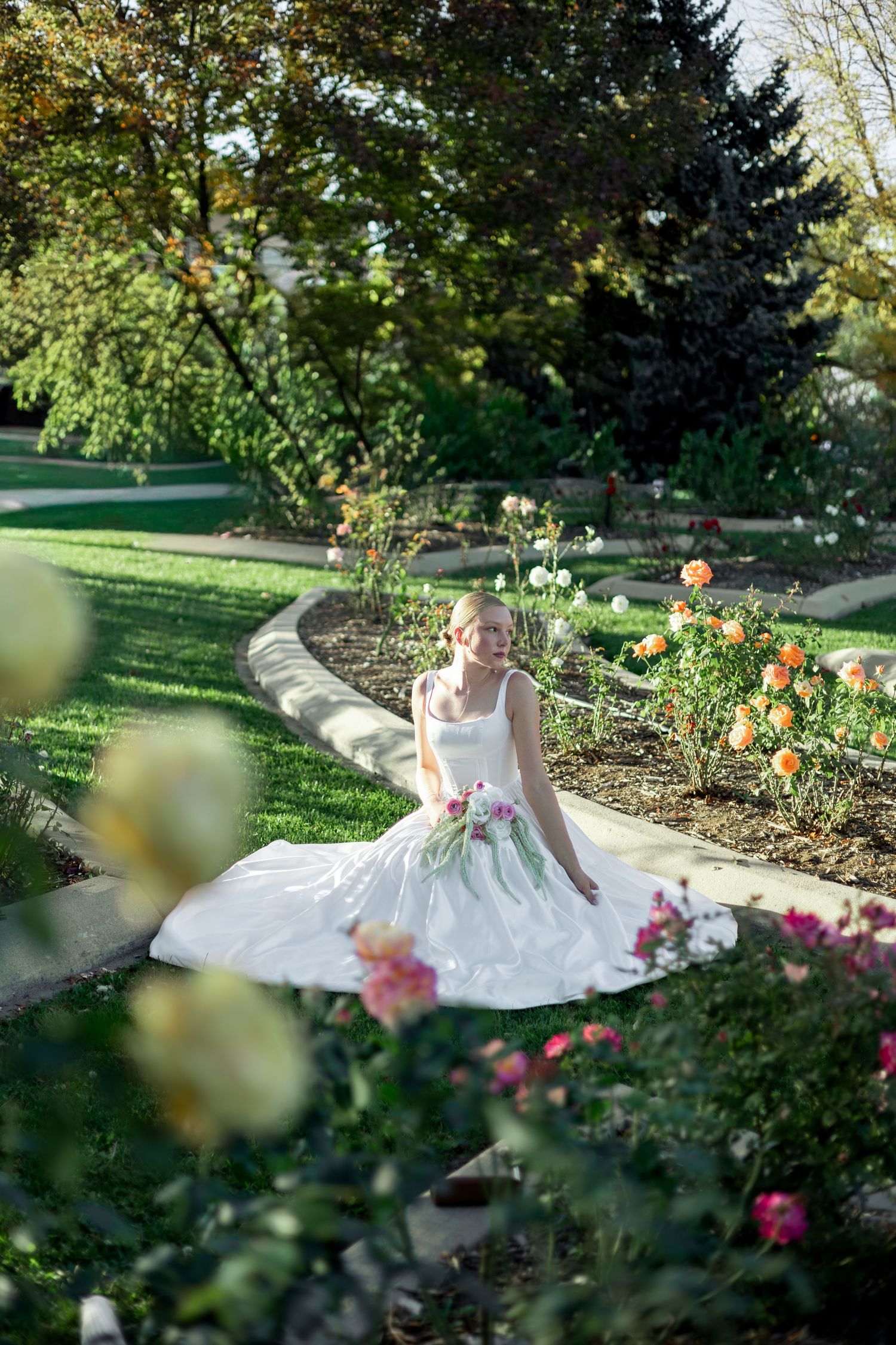 A bride in a white wedding dress poses gracefully among blooming flowers in a lush garden setting.