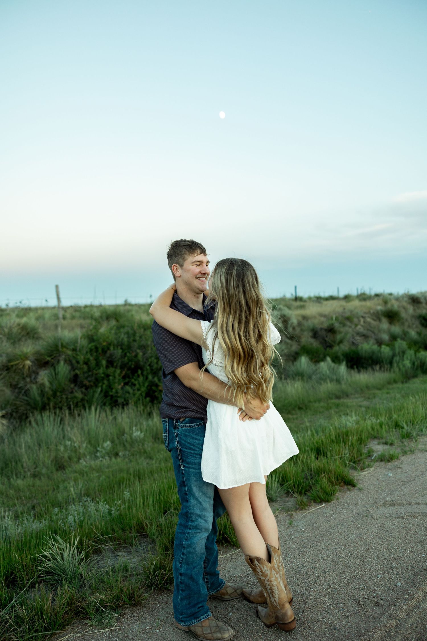 Couple embracing outdoors at dusk, woman in white dress and cowboy boots, man in jeans, green fields and blue sky behind them.