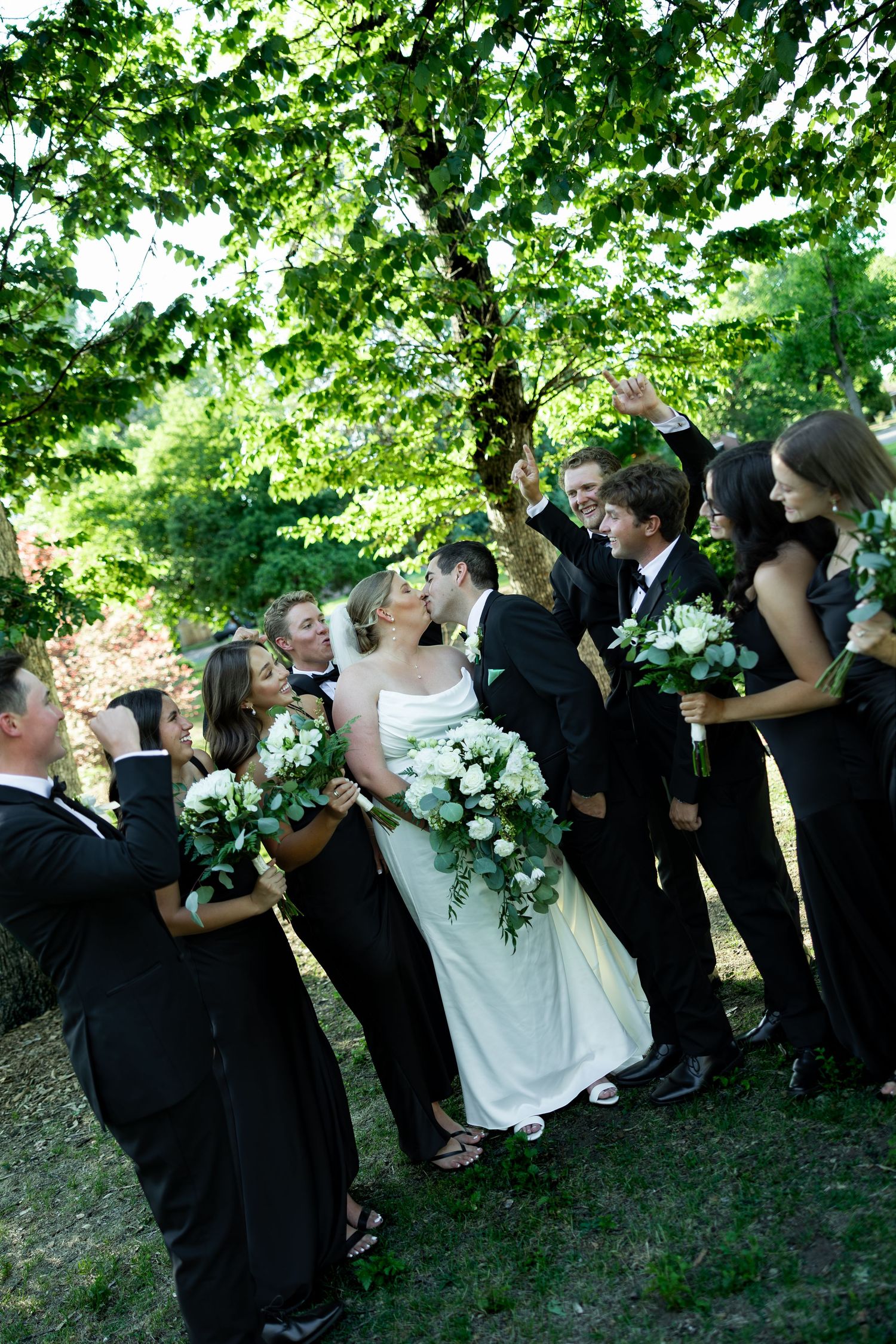 Wedding party in black attire surrounds kissing bride and groom outdoors under lush green trees.