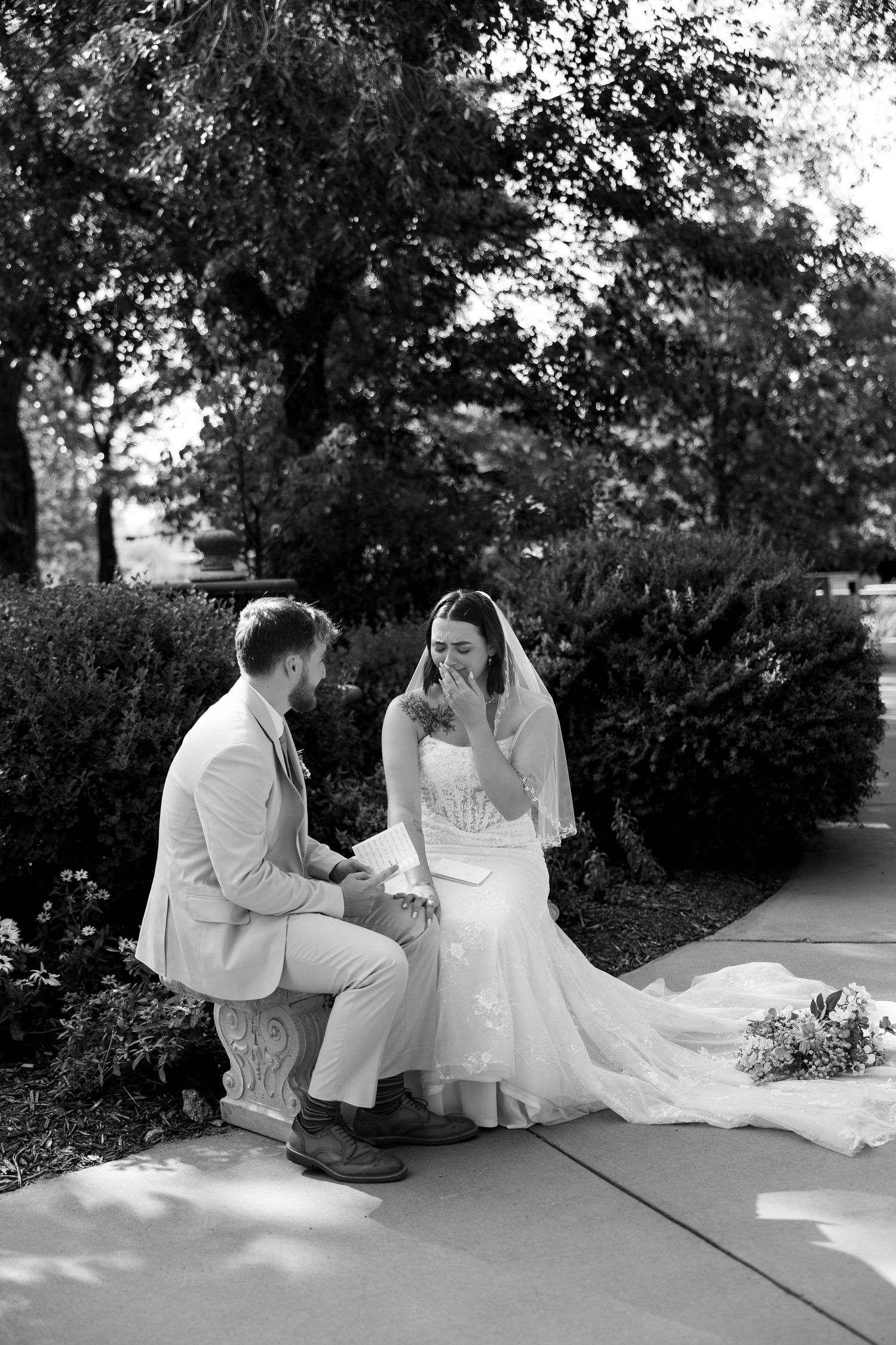 Black and white photo of groom kneeling before emotional crying bride in wedding dress outdoors.