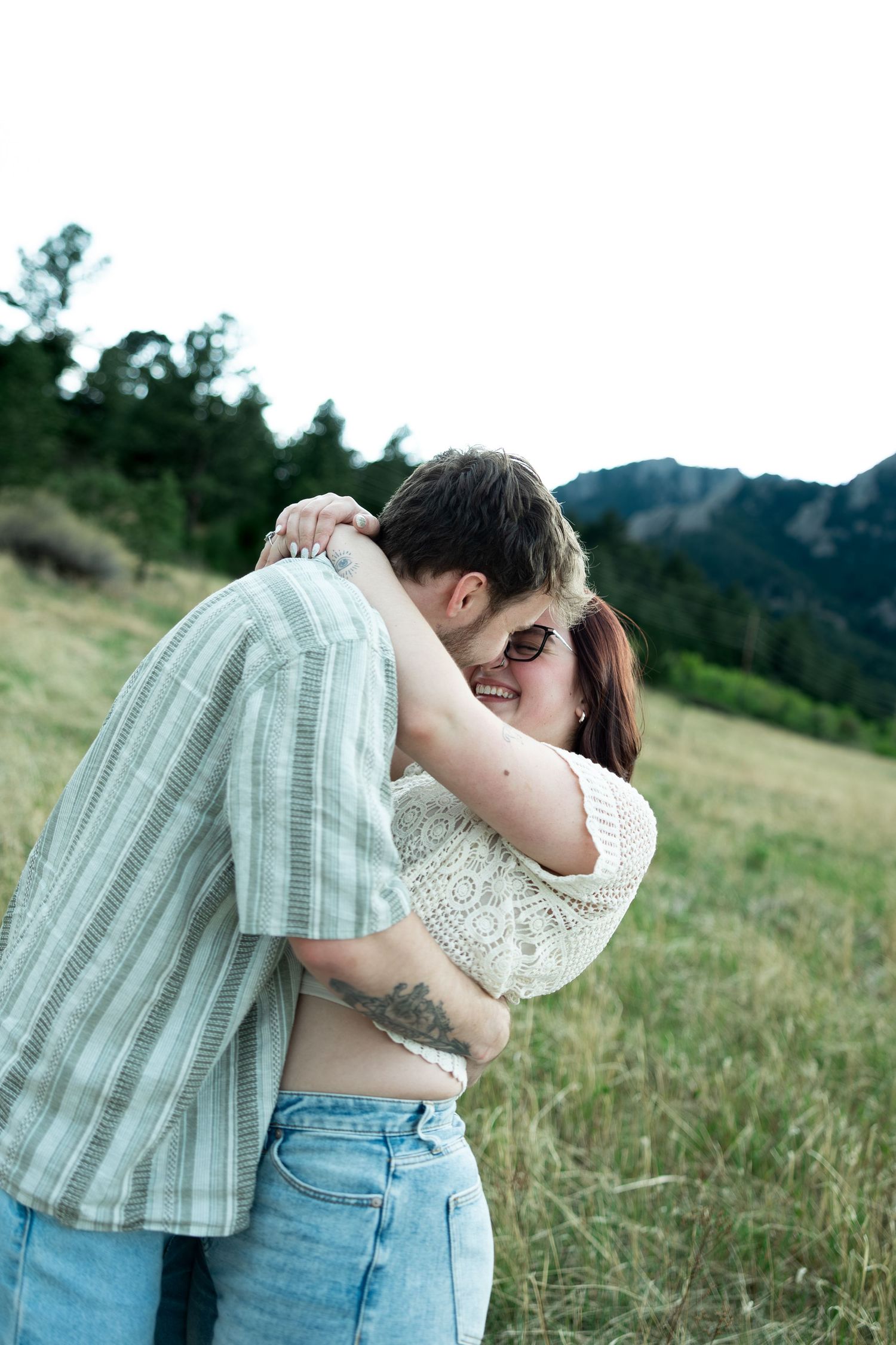 Couple embracing and laughing in a green mountain meadow, woman in lace top and jeans, man in plaid shirt.
