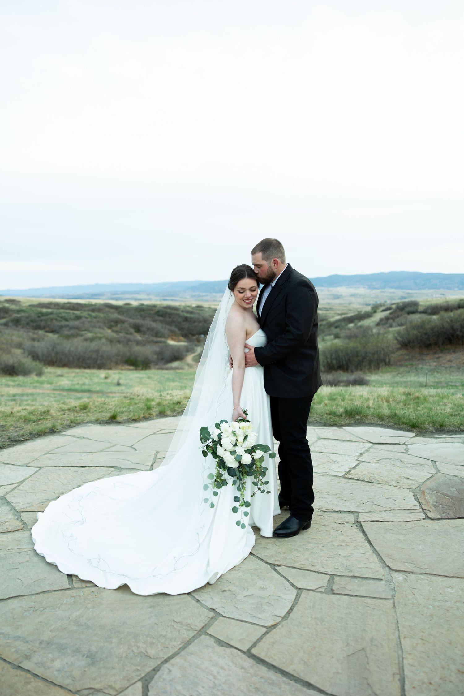 Bride in white gown with long veil holds bouquet as groom embraces her on stone terrace outdoors.