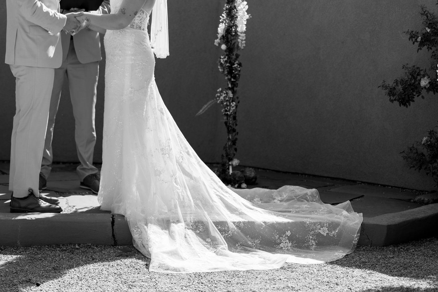 Black and white photo of a bride in a long lace wedding gown with train standing at outdoor ceremony.