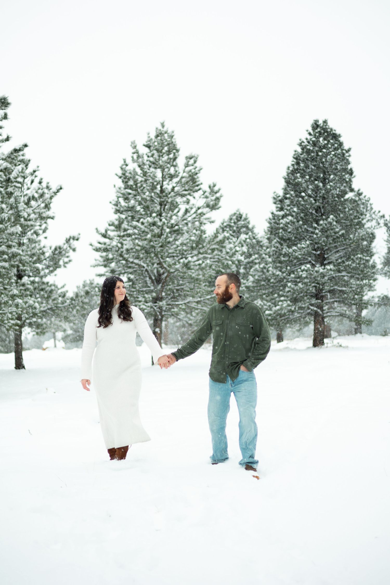 Couple holding hands in snowy winter landscape with snow-covered pine trees, woman in white dress and man in green shirt.