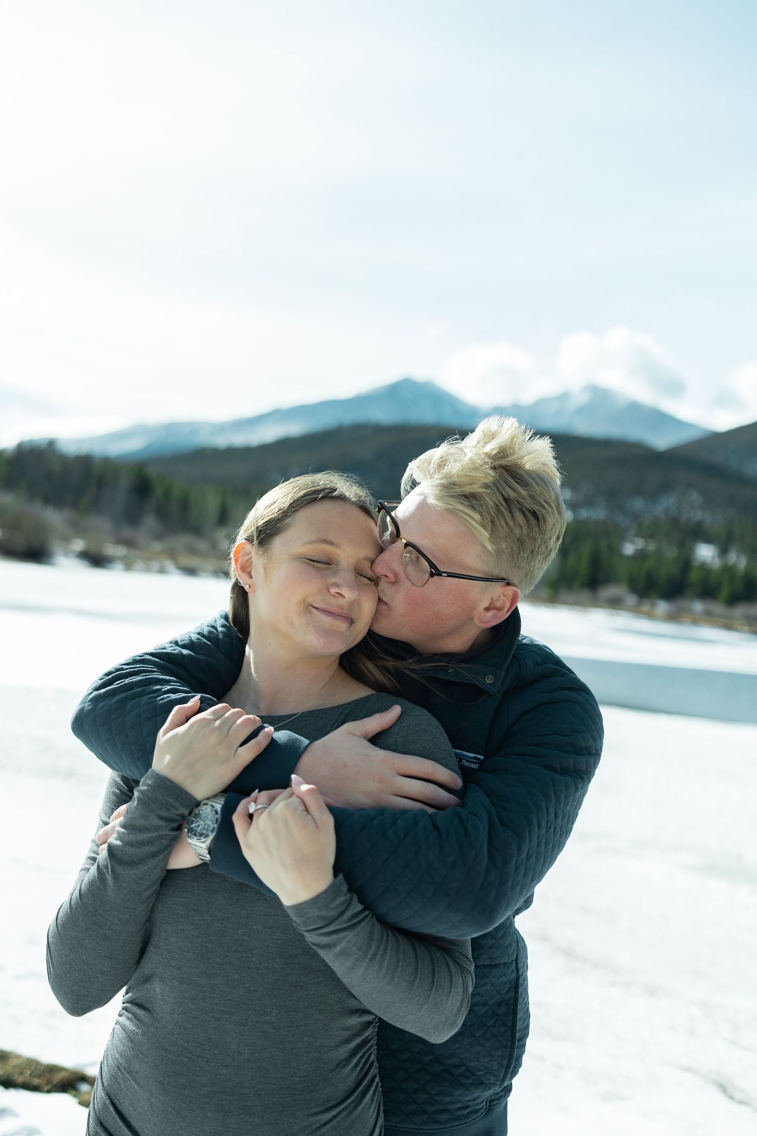 Couple embracing and kissing in snowy mountain landscape, woman smiling with eyes closed, wearing gray jacket.