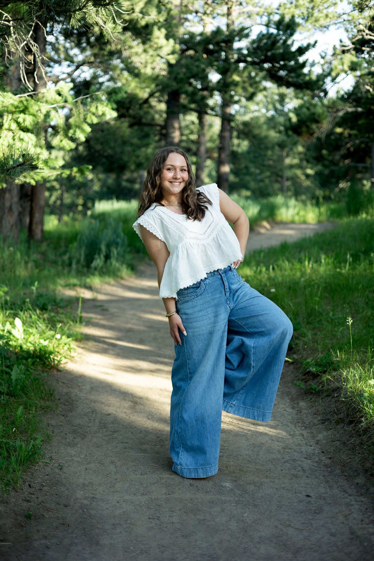A person in oversized denim baggy jeans and a white top poses playfully on a dirt path surrounded by greenery.