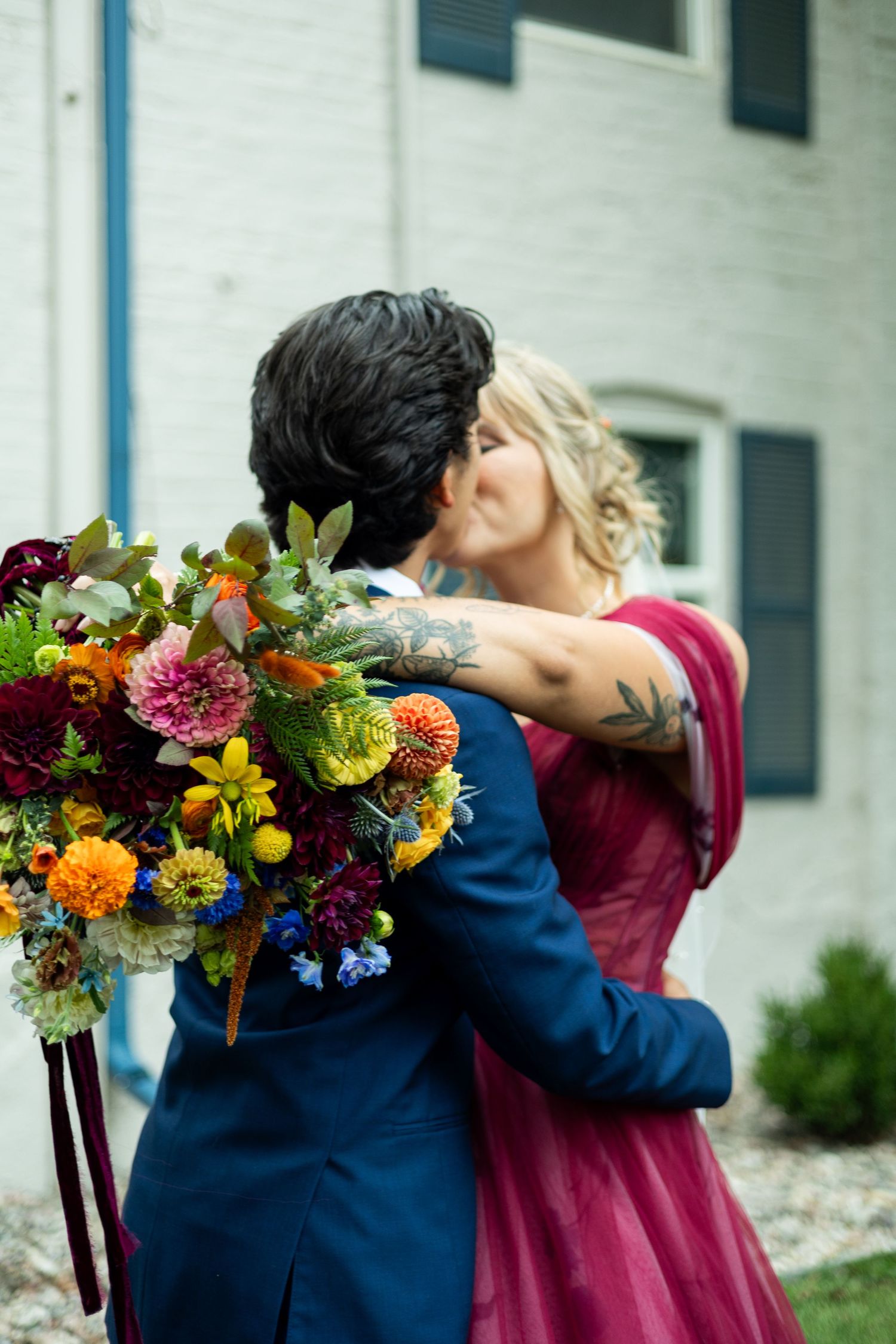 A couple embraces while holding a vibrant bouquet of wildflowers outside a white building.