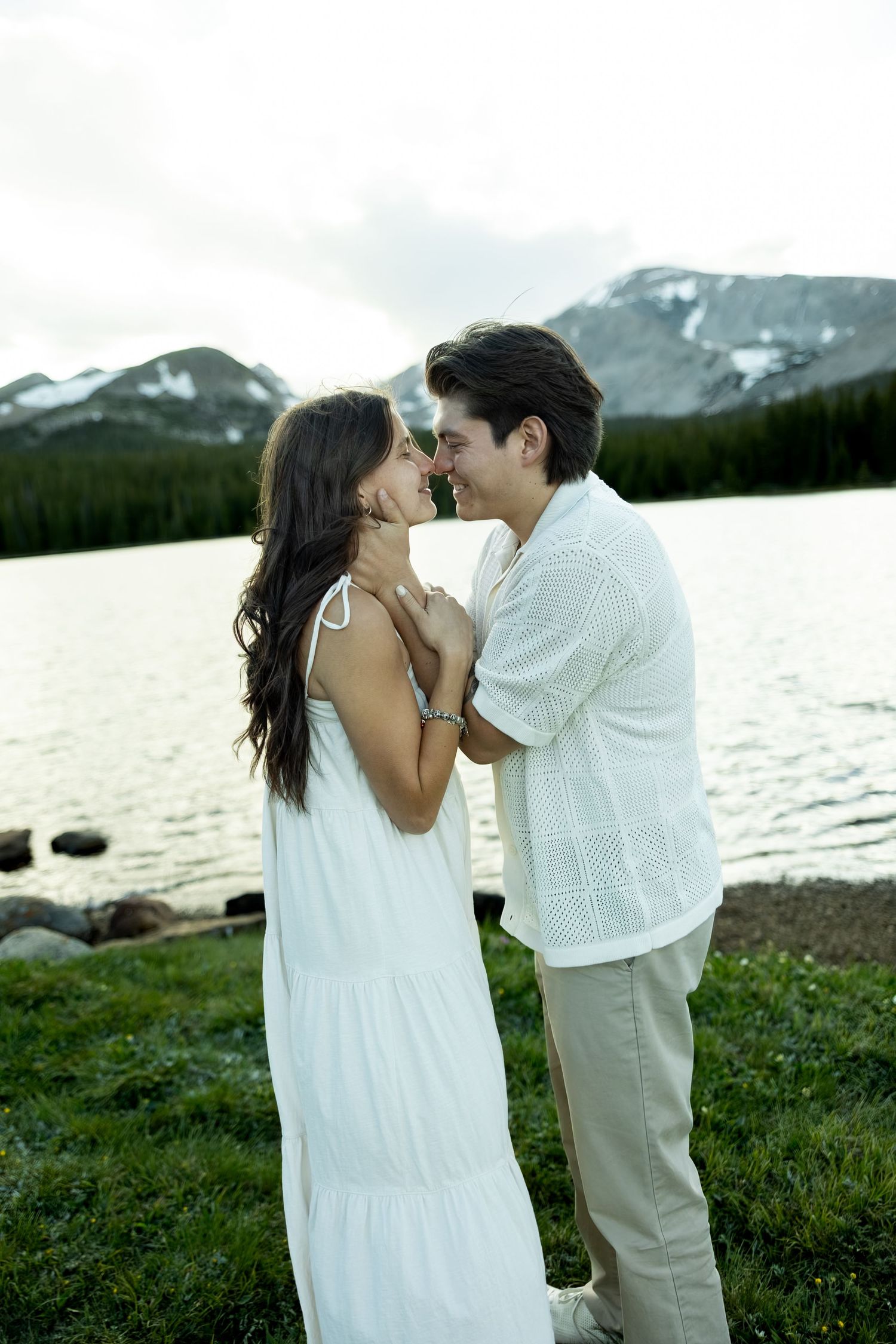 Romantic couple in white outfits sharing a tender moment by a mountain lake with snow-capped peaks.