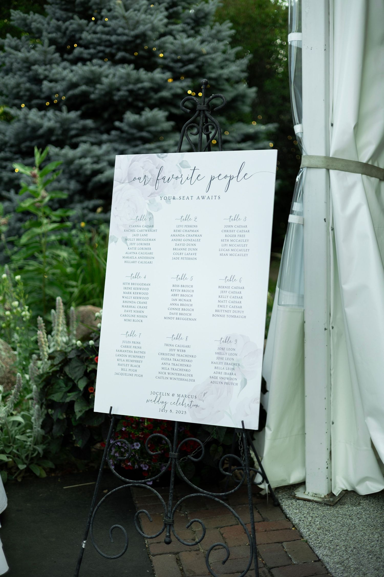 A wedding seating chart displayed on an ornate metal easel with blue spruce trees in the background.