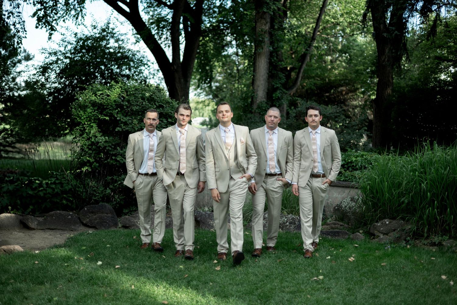 Groomsmen in light beige suits pose together in a garden setting.