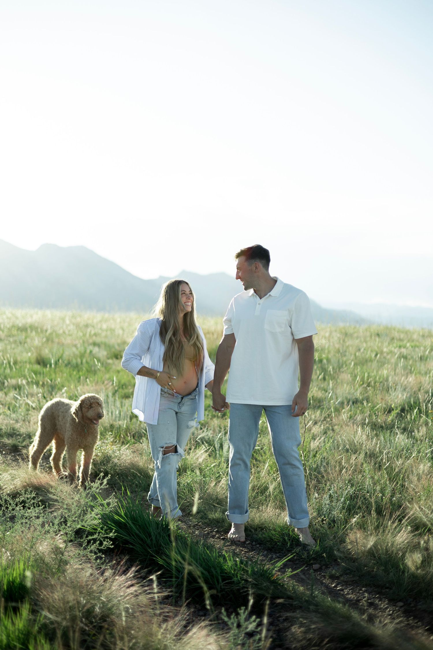 Two people in casual attire walking with their golden retriever on a scenic hillside during golden hour.