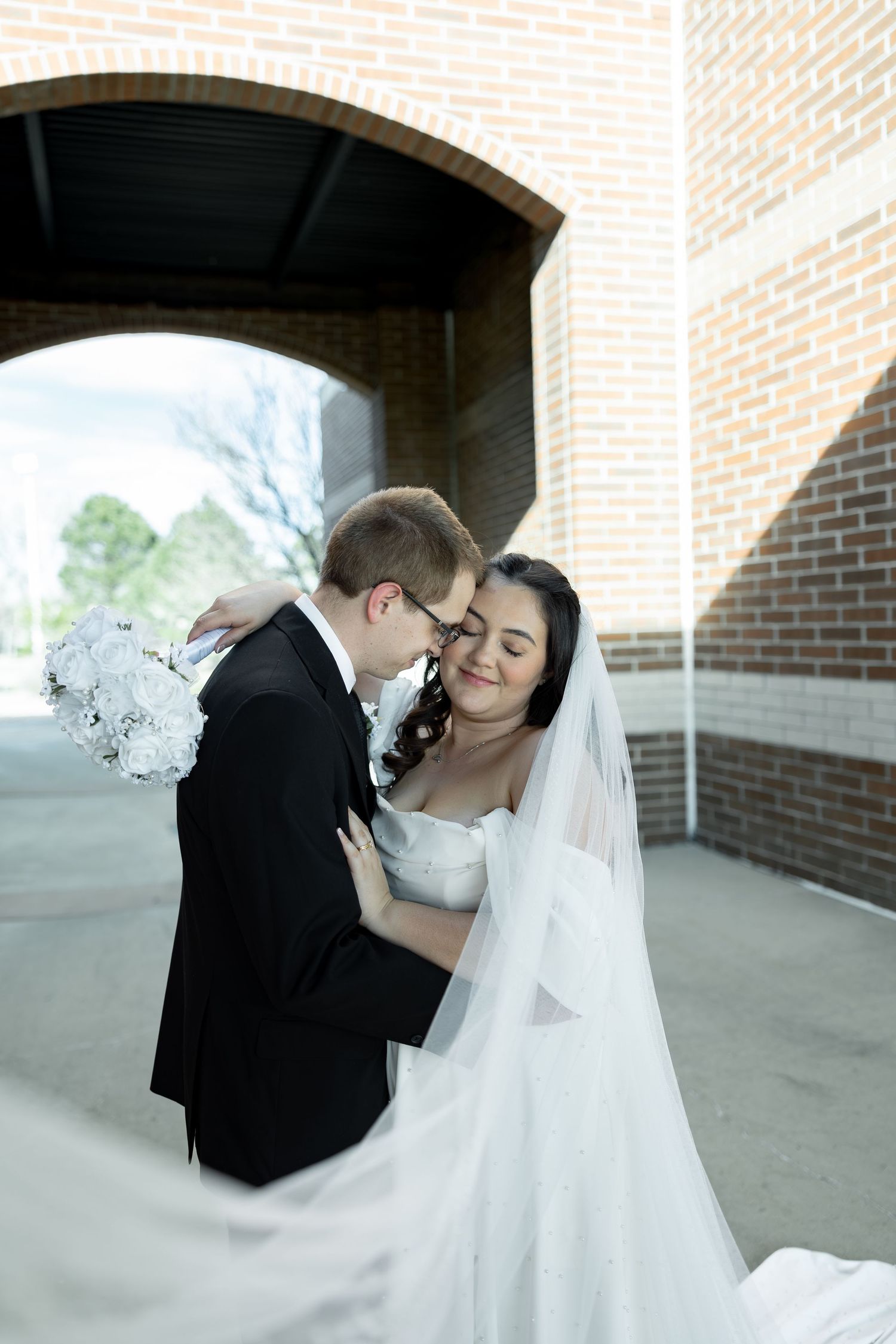 Bride and groom share a tender embrace under a brick archway, her white veil flowing gracefully.