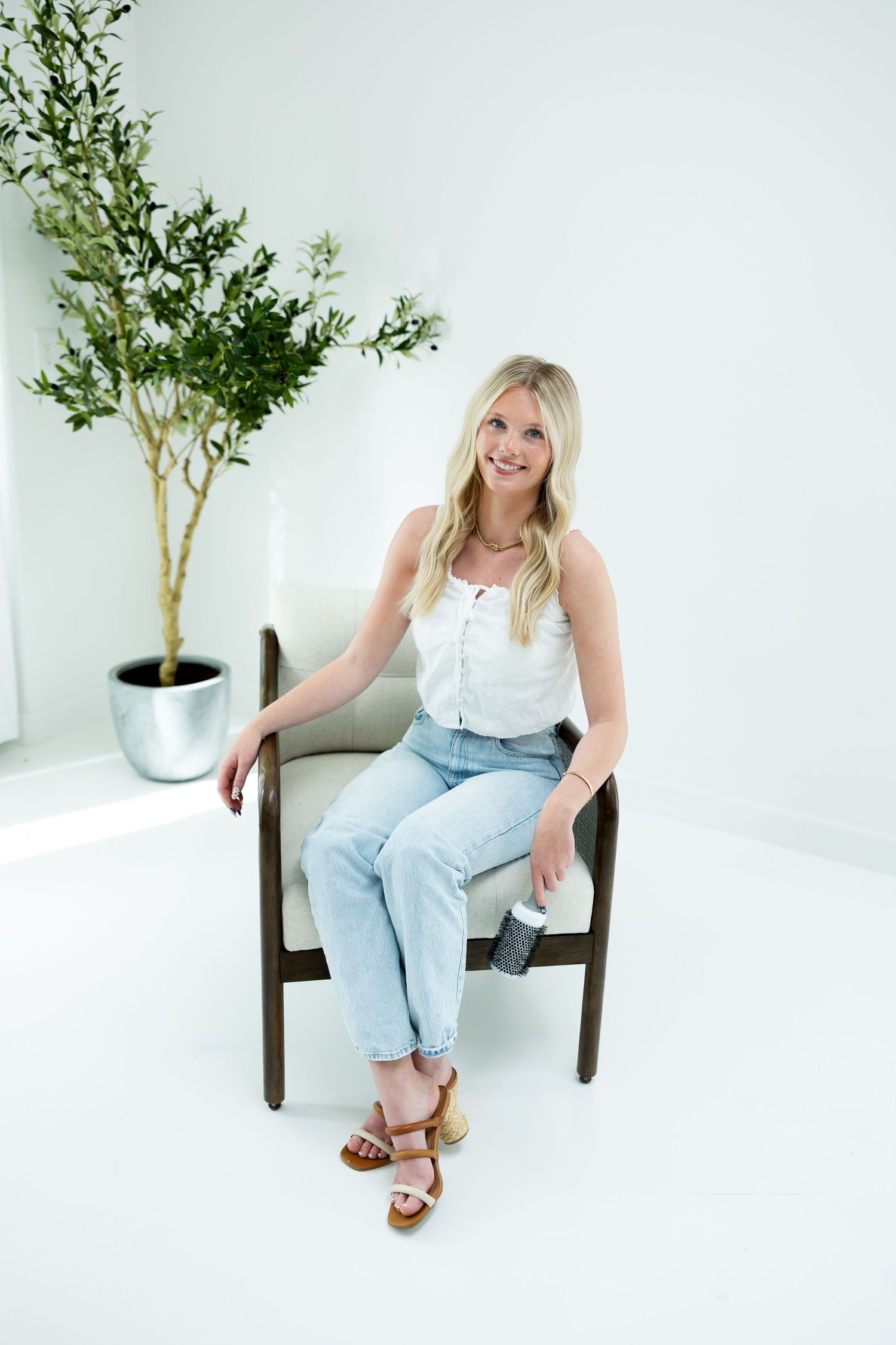 Smiling blonde woman in white top and jeans sitting in a modern chair beside a potted olive tree in a bright white room.
